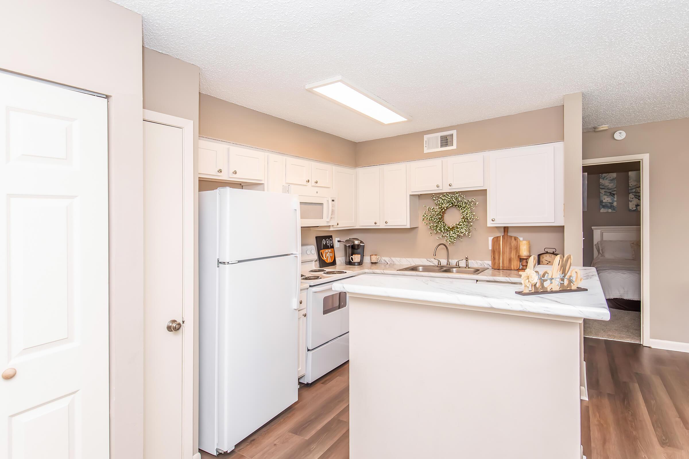 A modern kitchen featuring white cabinetry, a white refrigerator, and an electric stove. A small countertop island is visible with decorative items. The walls are painted in a neutral beige tone, and a wreath hangs on the wall. Natural light enters from the ceiling fixture, creating a bright atmosphere.