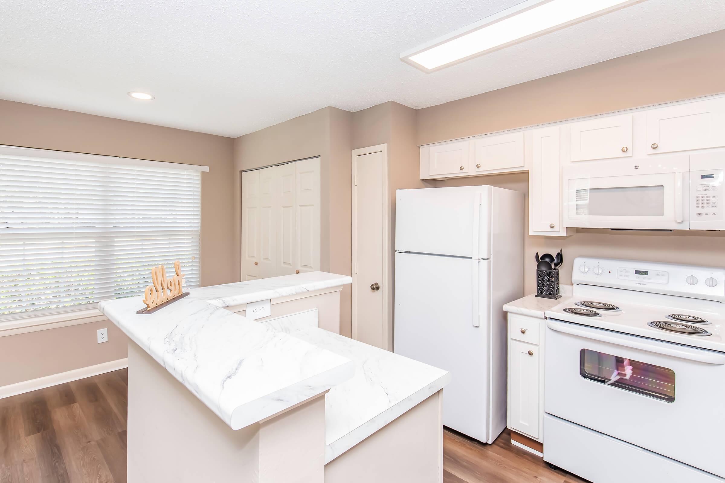 Bright modern kitchen featuring white cabinets, a marble countertop, and stainless steel appliances. The space includes a refrigerator, stove, and microwave, with natural light streaming in through a window. Neutral-colored walls create a warm and inviting atmosphere.
