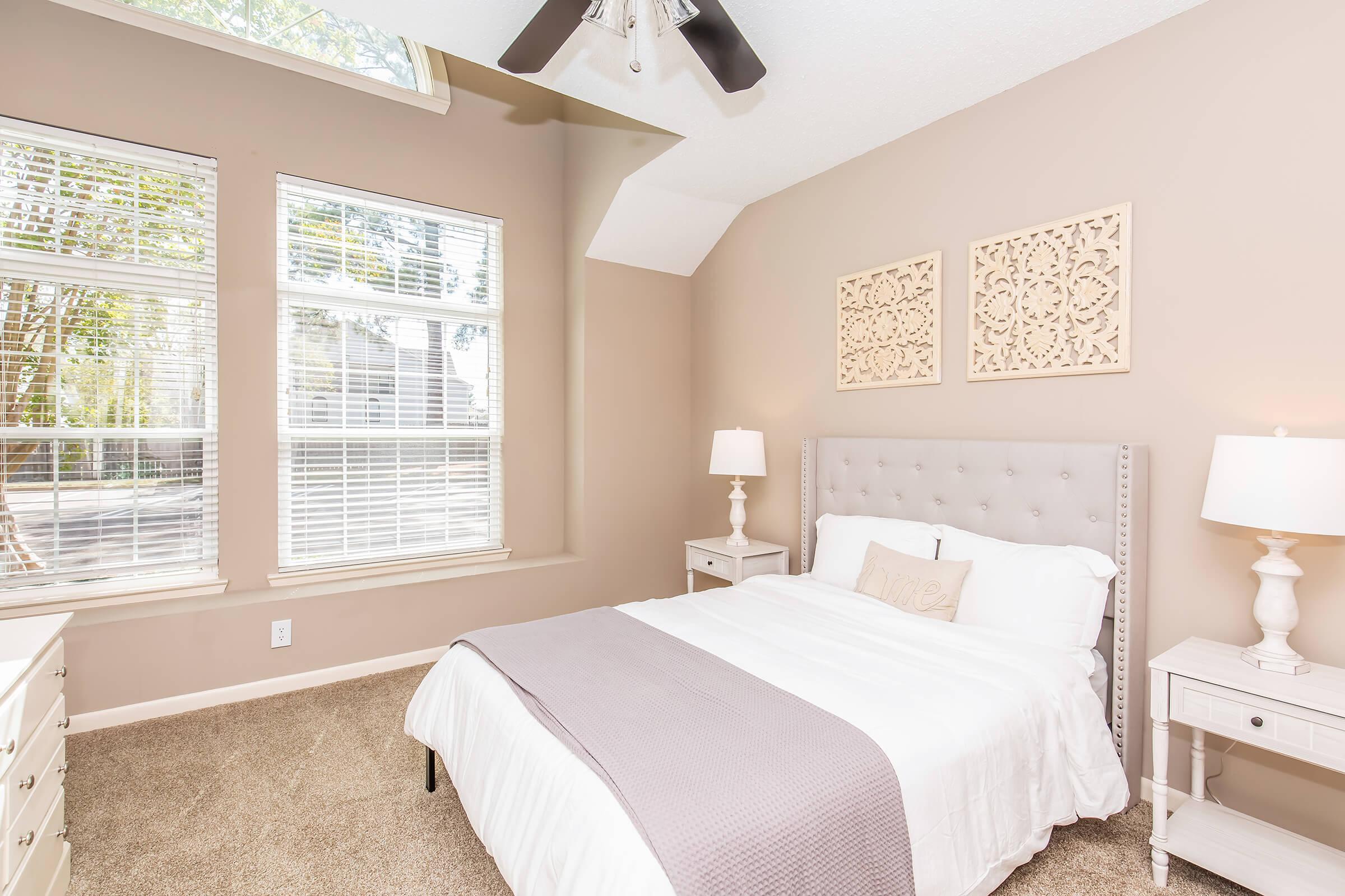 Bright and airy bedroom with a cream-colored bedspread on a tufted gray headboard. Two nightstands with lamps flank the bed. Soft, neutral walls complement large windows that allow natural light, and decorative wall art adds a touch of elegance. The carpeted floor enhances the cozy atmosphere.