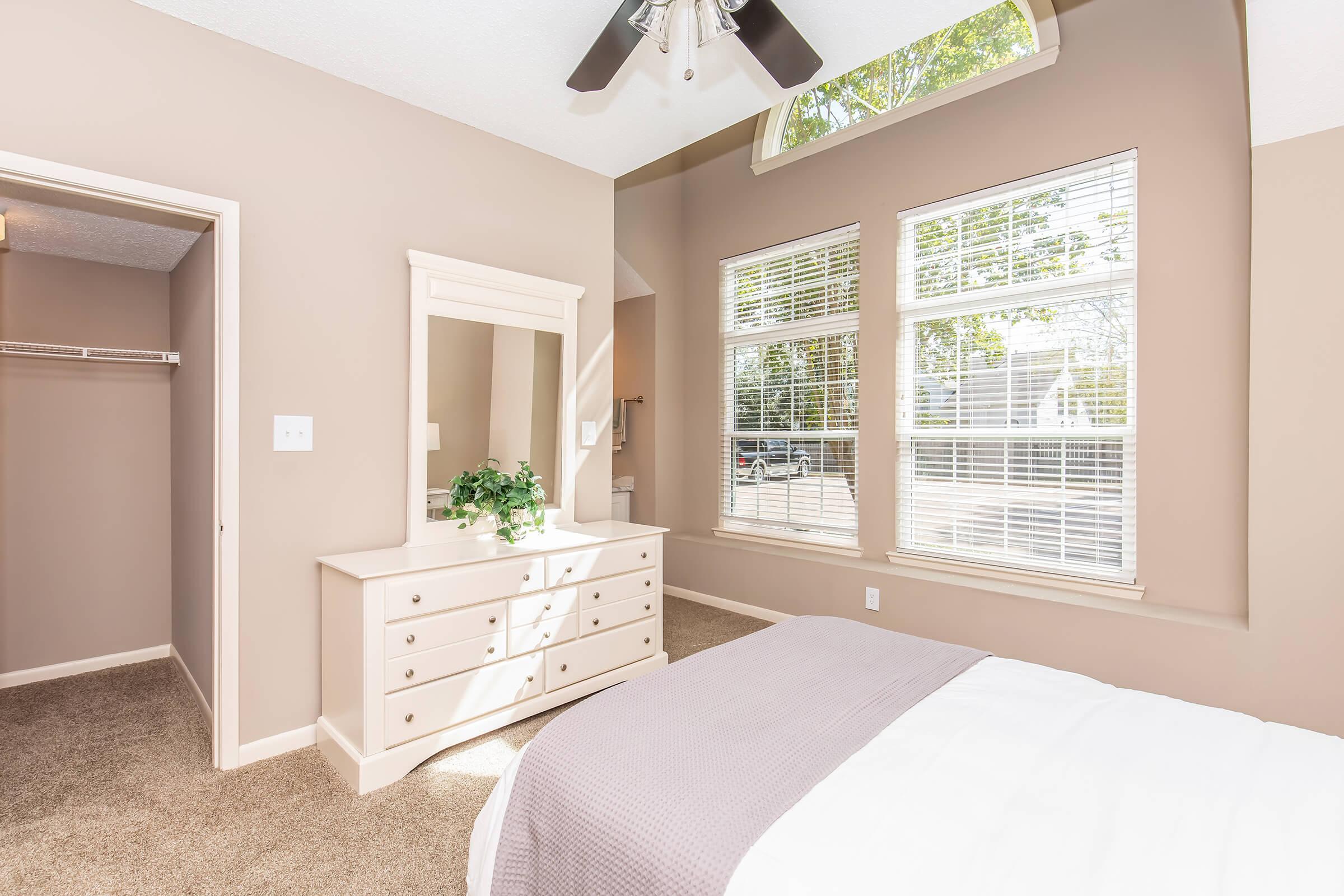 A cozy bedroom featuring light beige walls, a large window with ample natural light, a white dresser with a mirror, and a neatly made bed with a light-colored blanket. A closet is visible to the left, and the room has a plush carpet, creating a warm and inviting atmosphere.