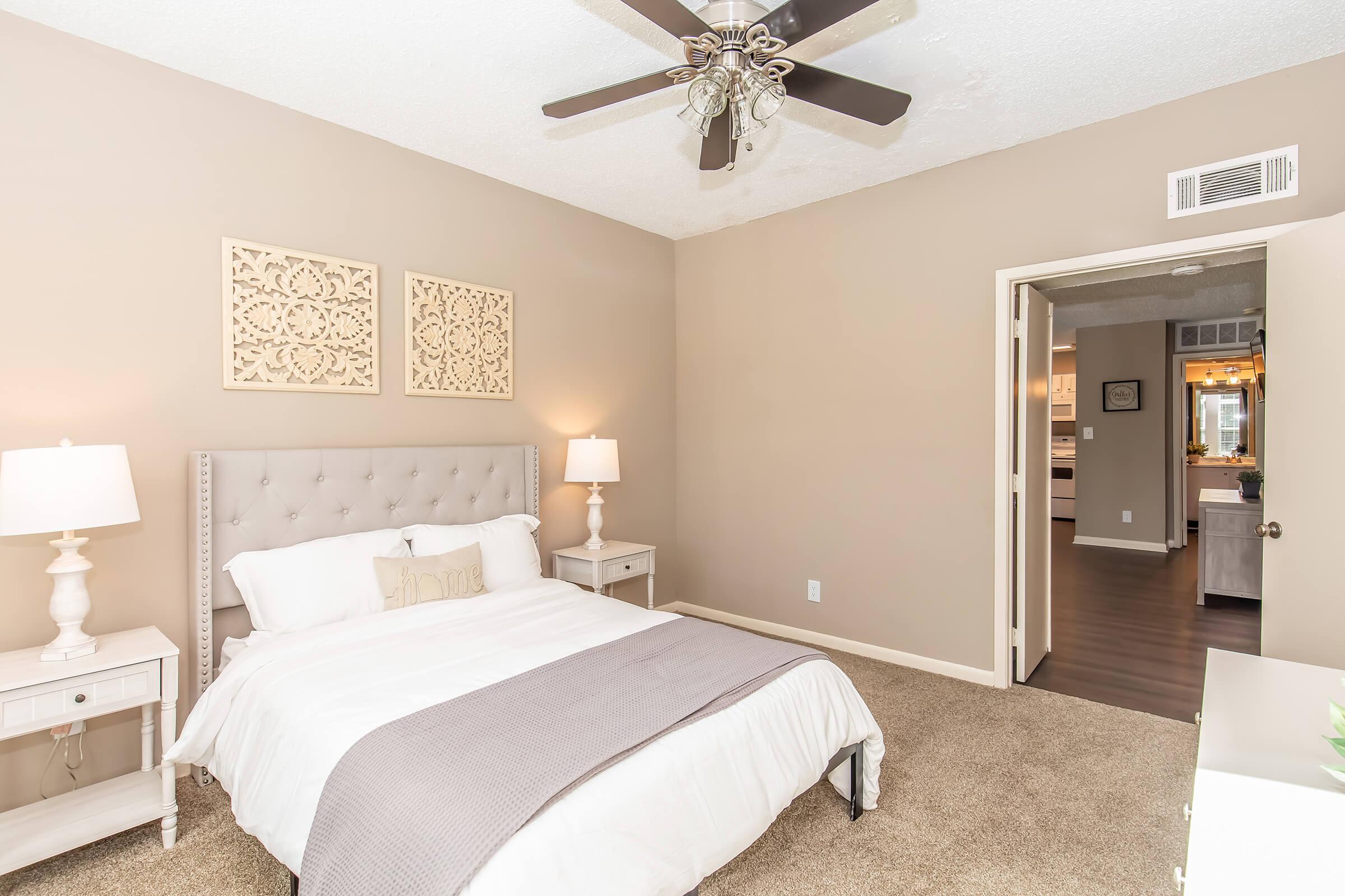 A cozy bedroom featuring a queen-sized bed with white bedding, a light gray throw blanket, and decorative pillows. Two bedside lamps on white nightstands illuminate the space. The walls are painted a soft beige, adorned with two artistic panels. A doorway leads to a kitchen area in the background.