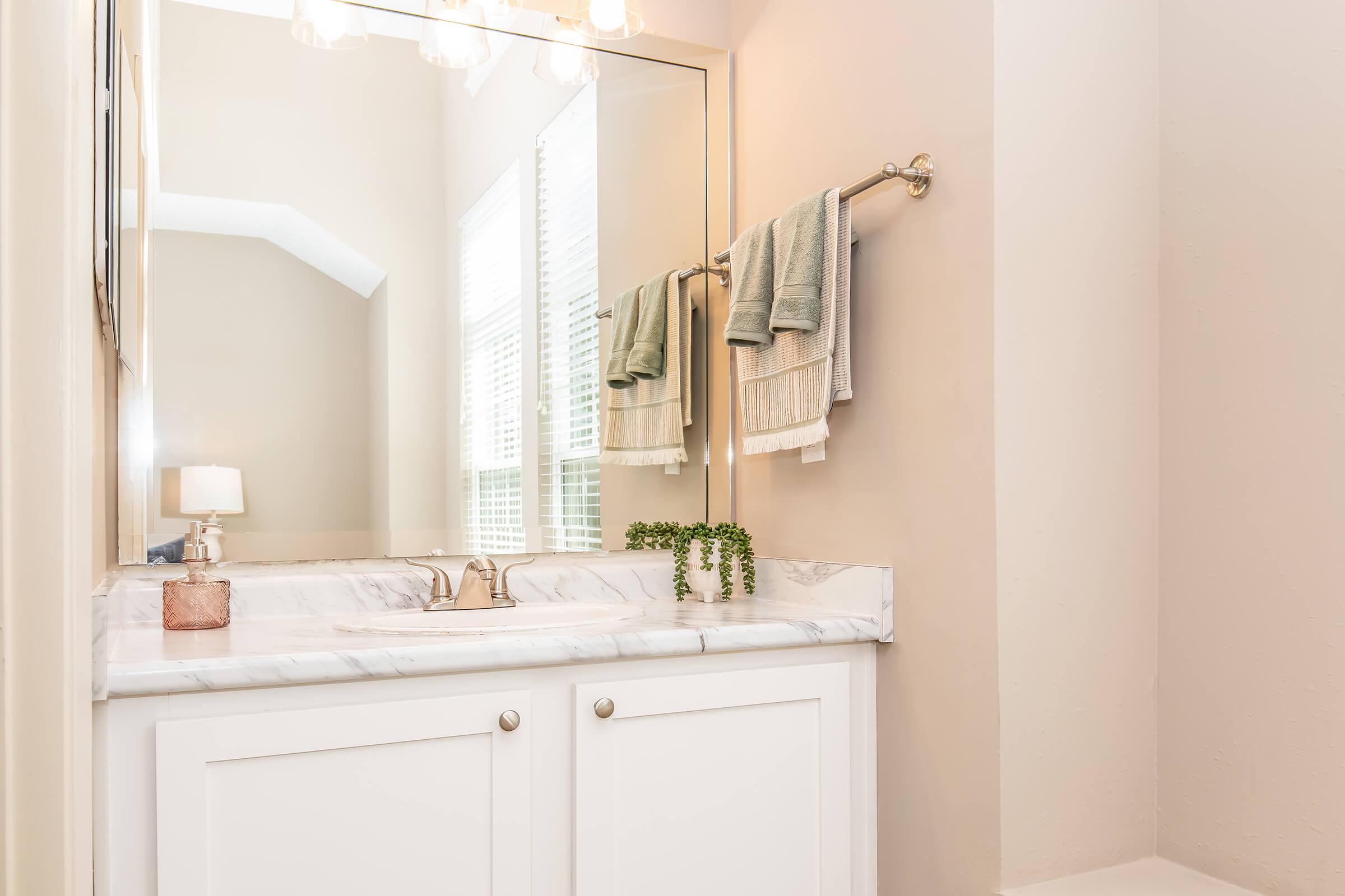 A modern bathroom featuring a white marble countertop with a gold faucet, a large mirror, and neatly arranged towels on a towel bar. Soft lighting illuminates the space, highlighting neutral wall colors and a small plant decoration on the counter. The overall look is clean and inviting.