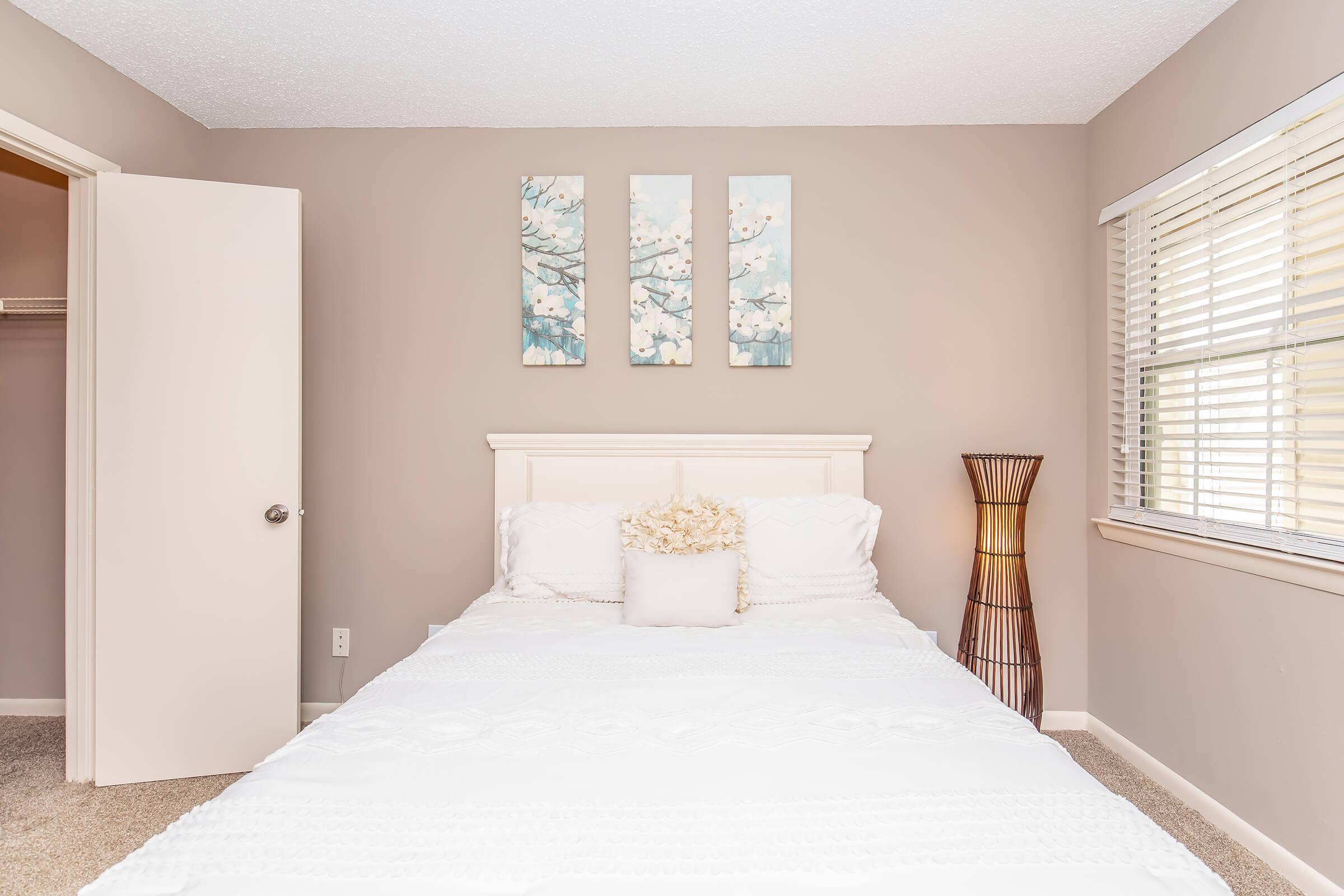 A cozy bedroom featuring a softly made bed with white bedding and decorative pillows, a tall wicker lamp beside it, and three framed art pieces depicting blossoming trees hanging on the light taupe wall. A closet door is partially open, and natural light filters through the window with blinds.