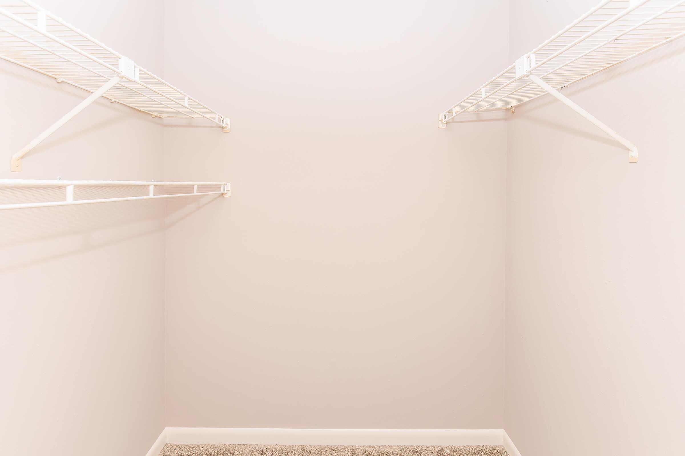 A spacious, empty closet with light beige walls and two white wire shelves on either side. The floor is covered with a light-colored carpet, and the overall ambiance is clean and minimalistic, ideal for organizing clothes and items.