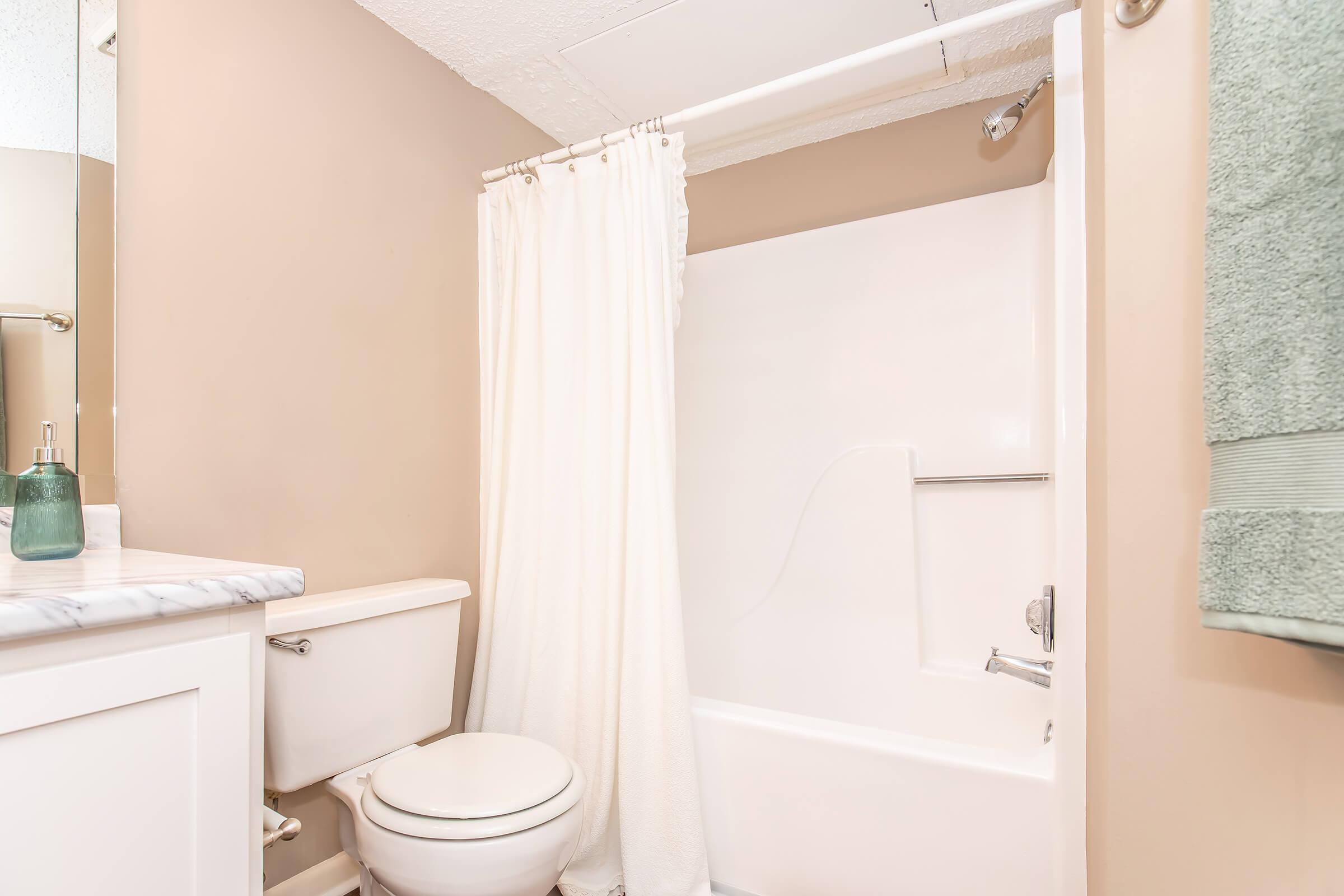 A clean and minimalistic bathroom featuring a white bathtub with a shower curtain, a white toilet, and a small sink with a marble countertop. The walls are painted a soft beige color, and there is a green towel hanging nearby. The space is well-lit and tidy, creating a fresh atmosphere.