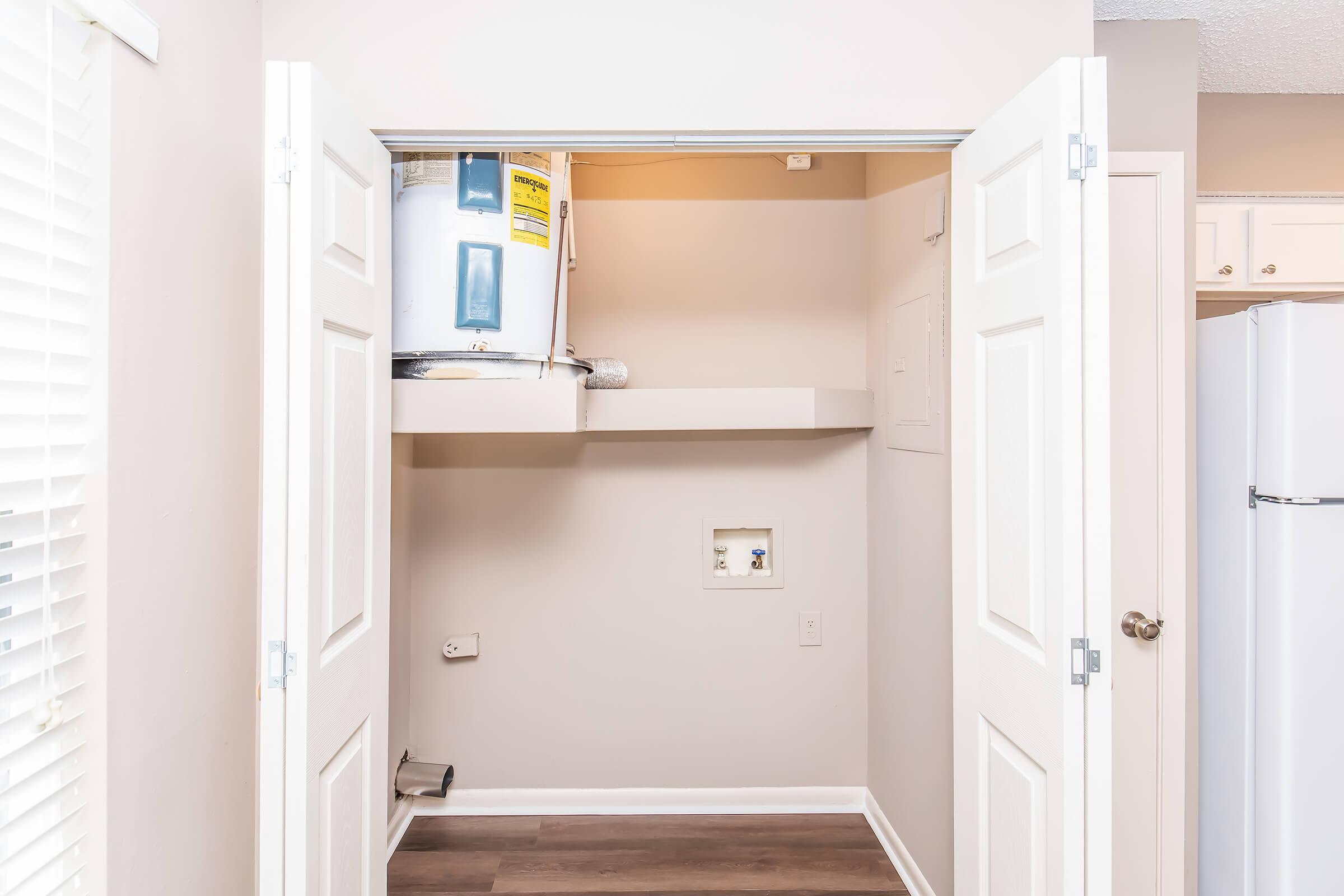 A small laundry closet with two white doors opened, revealing a water heater mounted on the wall and a shelf above. The closet features a power outlet on the wall and a small space below the water heater. Light beige walls complement the wooden flooring.