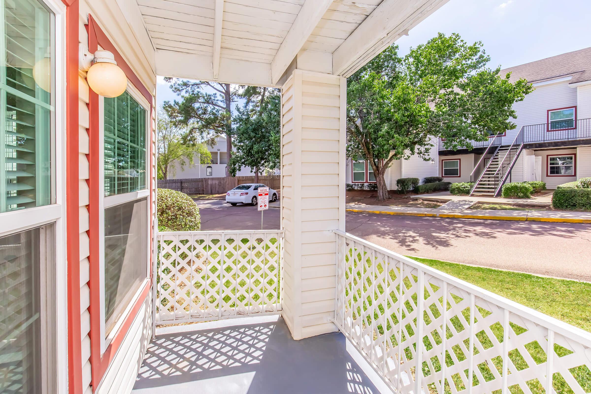 View from a covered porch featuring white lattice railings. The scene includes a well-maintained lawn, trees, and a parking area with parked cars in the background. The sky is clear, indicating a sunny day. The setting appears to be part of a residential complex.