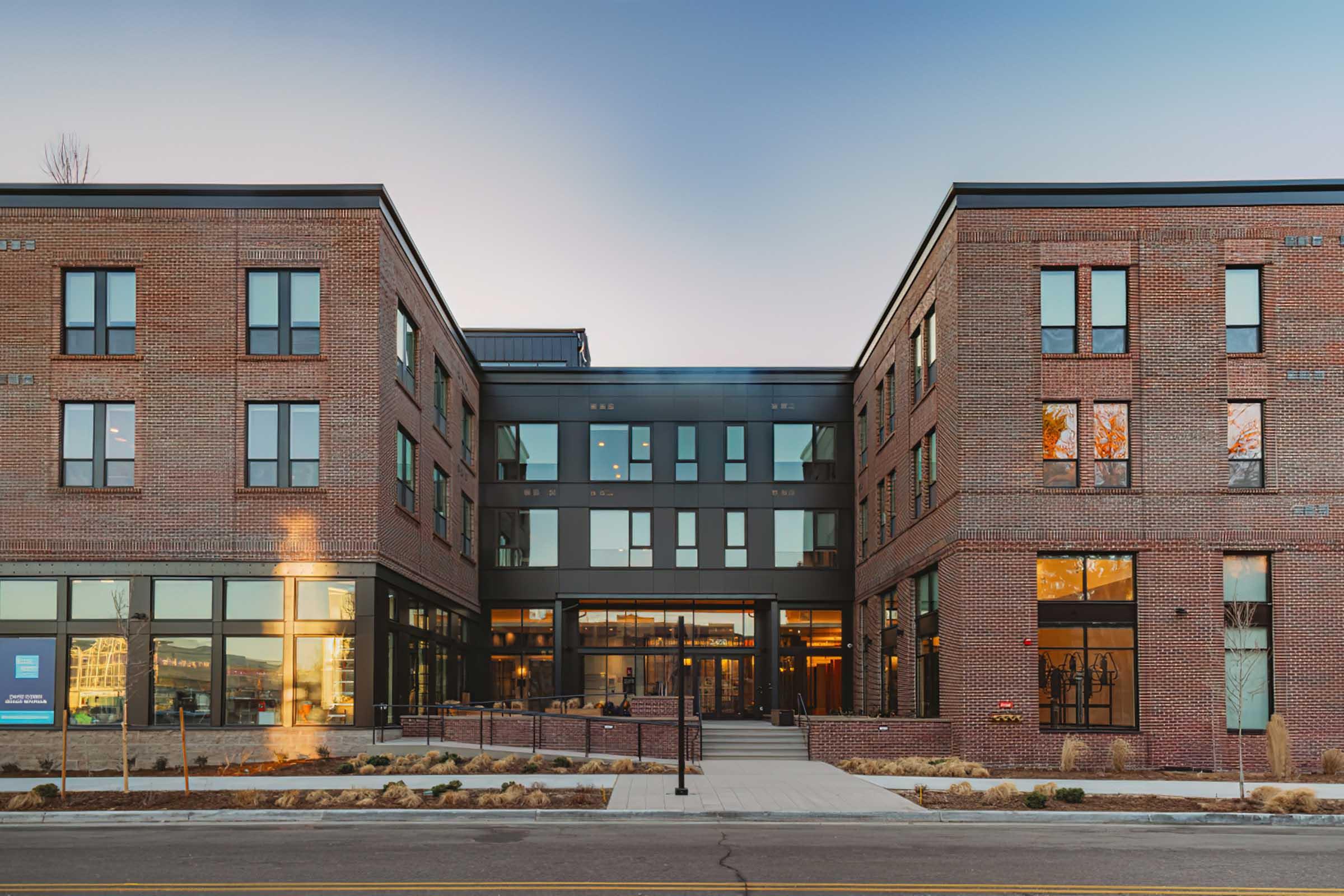 Modern brick building with large windows, featuring a mix of industrial and contemporary design elements. The structure has two wings flanking a central entrance, surrounded by landscaping and walkways, under a clear blue sky. The scene conveys a welcoming atmosphere with a stylish architectural aesthetic.