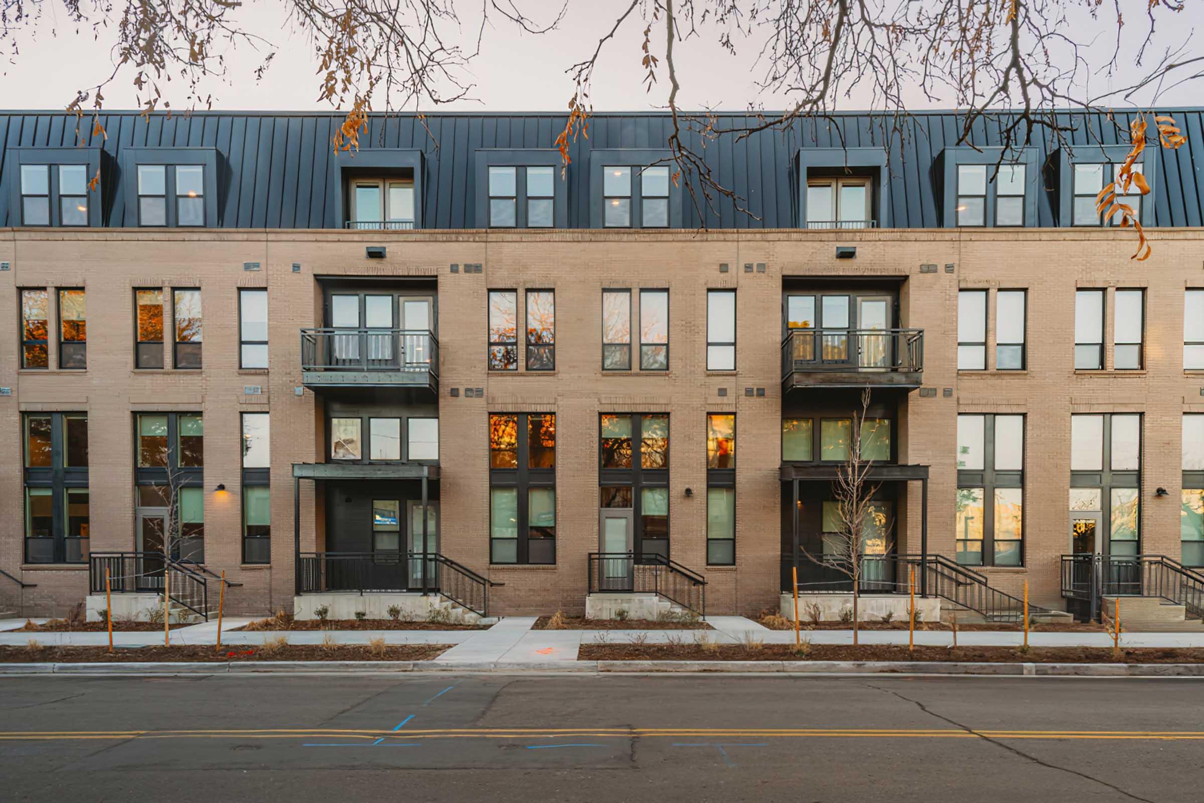 A modern multi-unit residential building featuring a brick facade with large windows and balconies. The structure has a flat roof and is set along a street with a sidewalk. Surrounding trees are visible, and the scene captures warm reflections in the windows, suggesting a late afternoon or early evening setting.