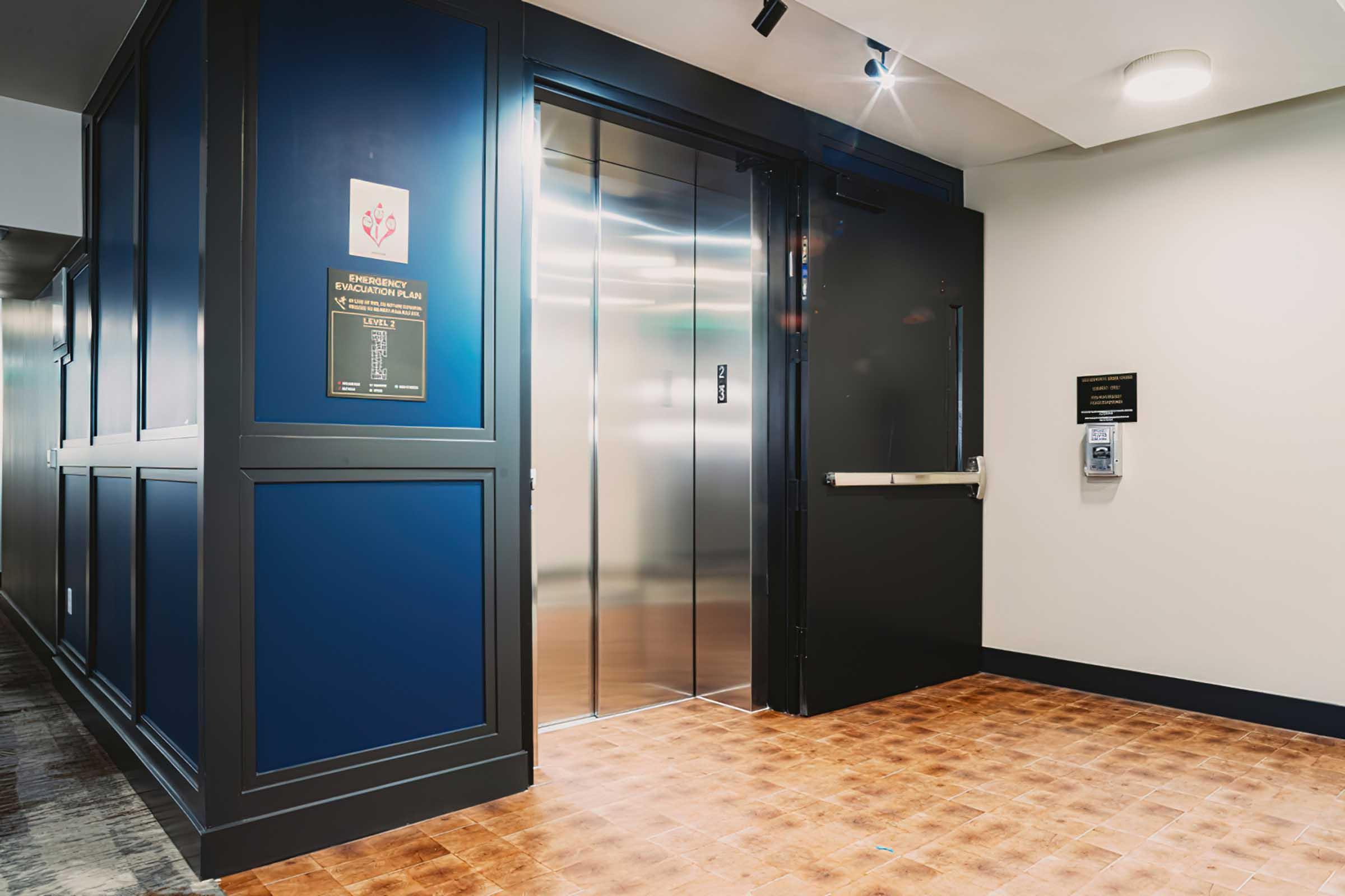 Elevator entrance in a modern building, featuring a polished metal door and blue paneling. Emergency information is displayed on a sign beside the elevator. The floor is tiled in a warm, wood-like pattern, with recessed lighting illuminating the space.