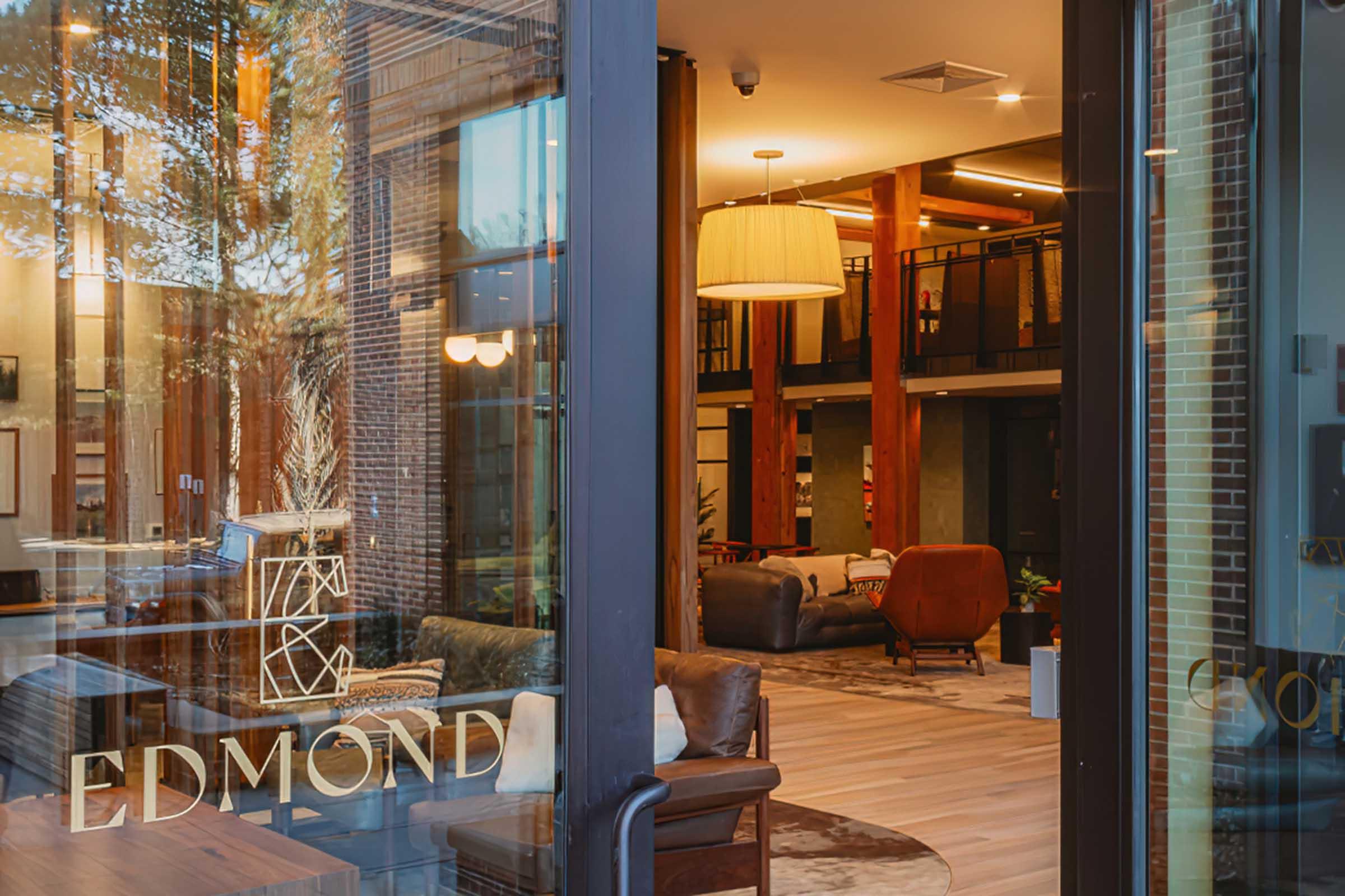 View of a stylish lobby area through glass doors, featuring modern furnishings like chairs and sofas, warm lighting, and wooden accents. The logo "EDMOND" is prominently displayed on the door. The space has a welcoming and contemporary ambiance, with trees visible outside.