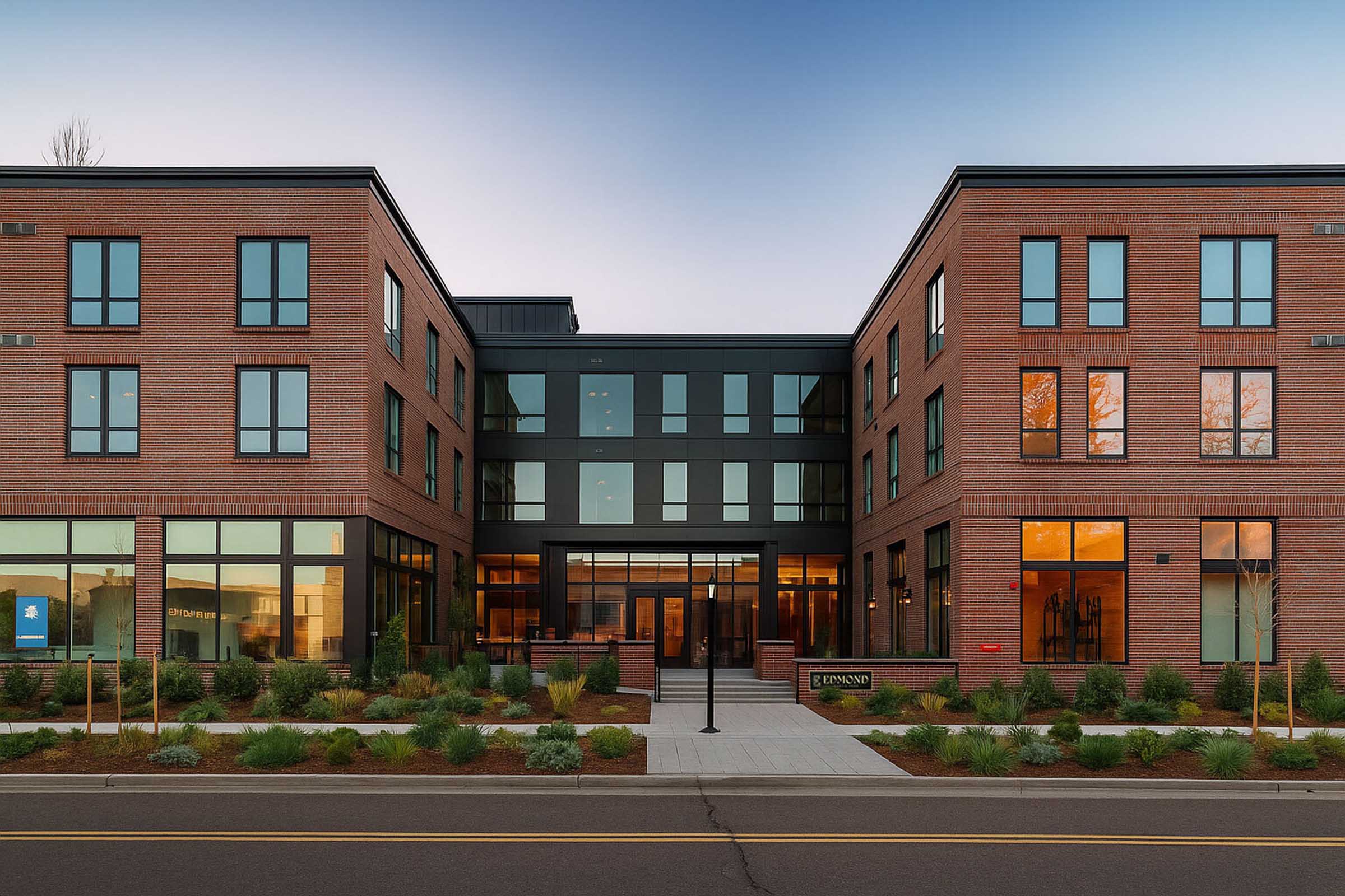 Modern brick building with large windows, featuring a central entrance framed by two symmetrical wings. The facade includes a mix of glass and brick elements, surrounded by landscaped greenery. The scene is set during twilight, with warm light glowing from the windows, creating an inviting atmosphere.
