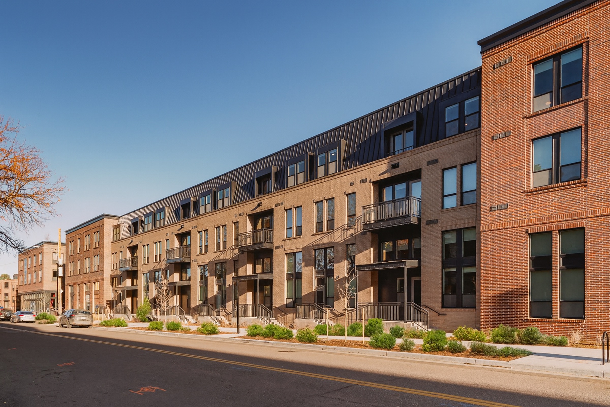 Row of modern multi-story apartment buildings with a mix of brick and metal finishes, featuring balconies and large windows. The street in front is lined with small shrubs and trees, and parked cars are visible along the curb. The sky above is clear and blue, indicating a sunny day.