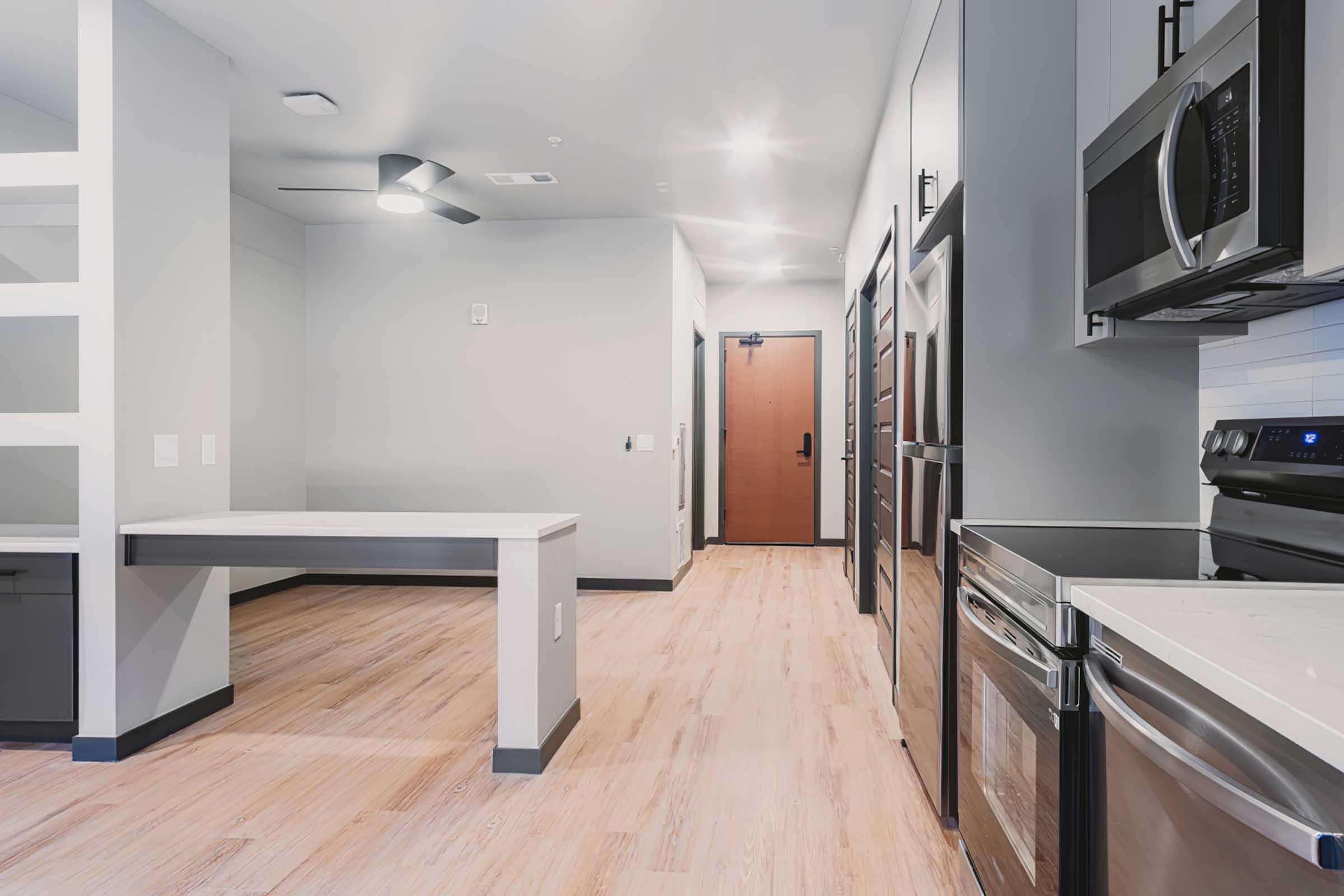A modern kitchen and dining area featuring stainless steel appliances, a spacious countertop, and wood-like flooring. The layout includes a door leading outside, a ceiling fan, and neutral-colored walls with minimalistic design elements.