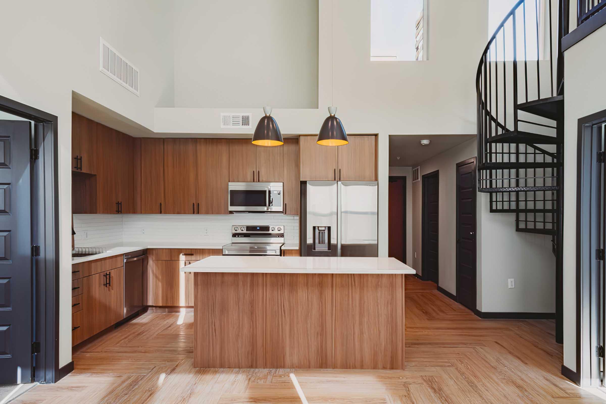 Modern kitchen with wooden cabinets and a spacious island featuring a white countertop. Includes stainless steel appliances, a microwave, and a spiral staircase leading to an upper level. The design is sleek and contemporary, with pendant lighting and dark doors contrasting against light walls.