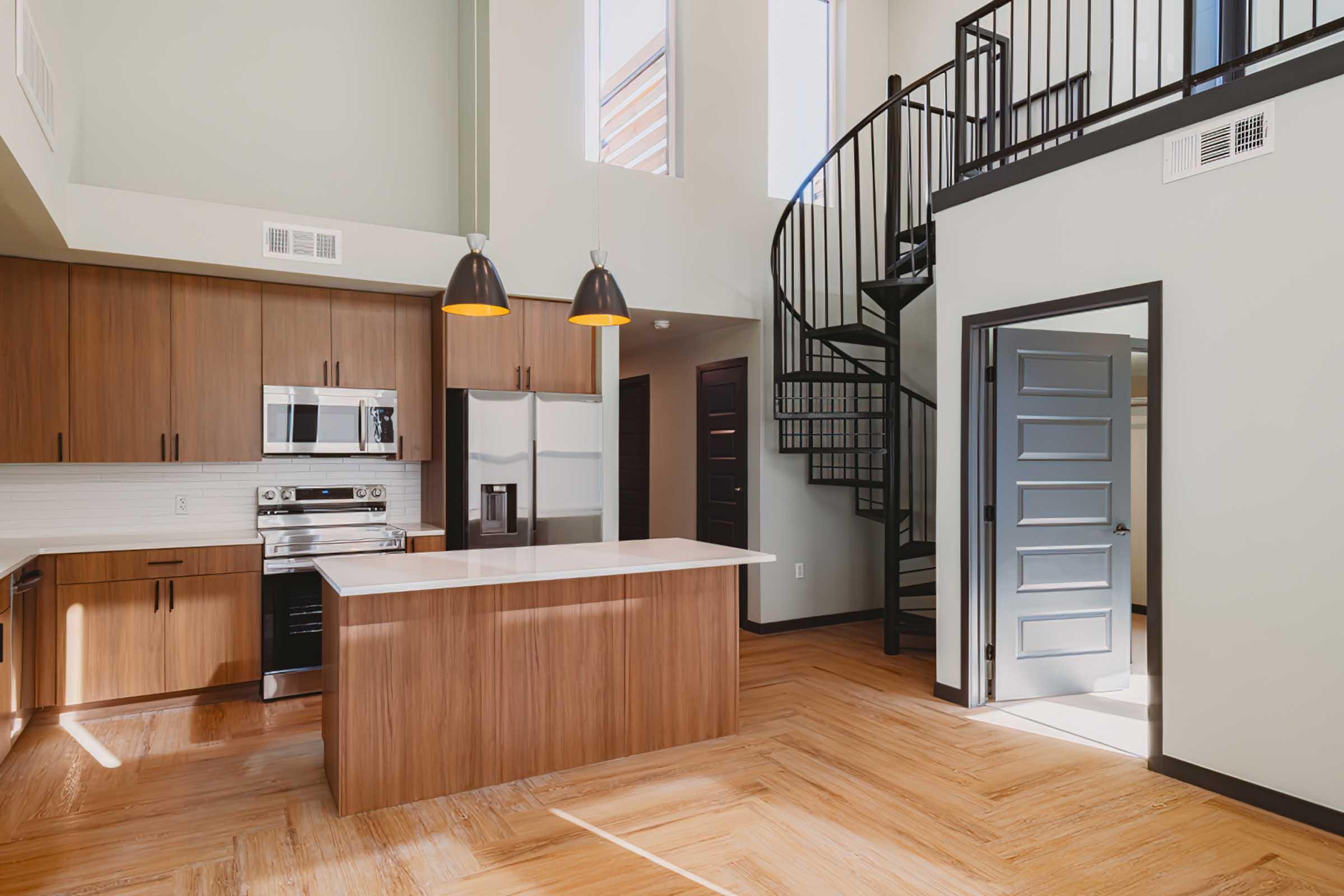 Modern kitchen and living area featuring wooden cabinets, stainless steel appliances, and a white countertop island. A spiral staircase leads to an upper level, with large windows allowing natural light to brighten the space. The floor is made of light-colored wood with a herringbone pattern.
