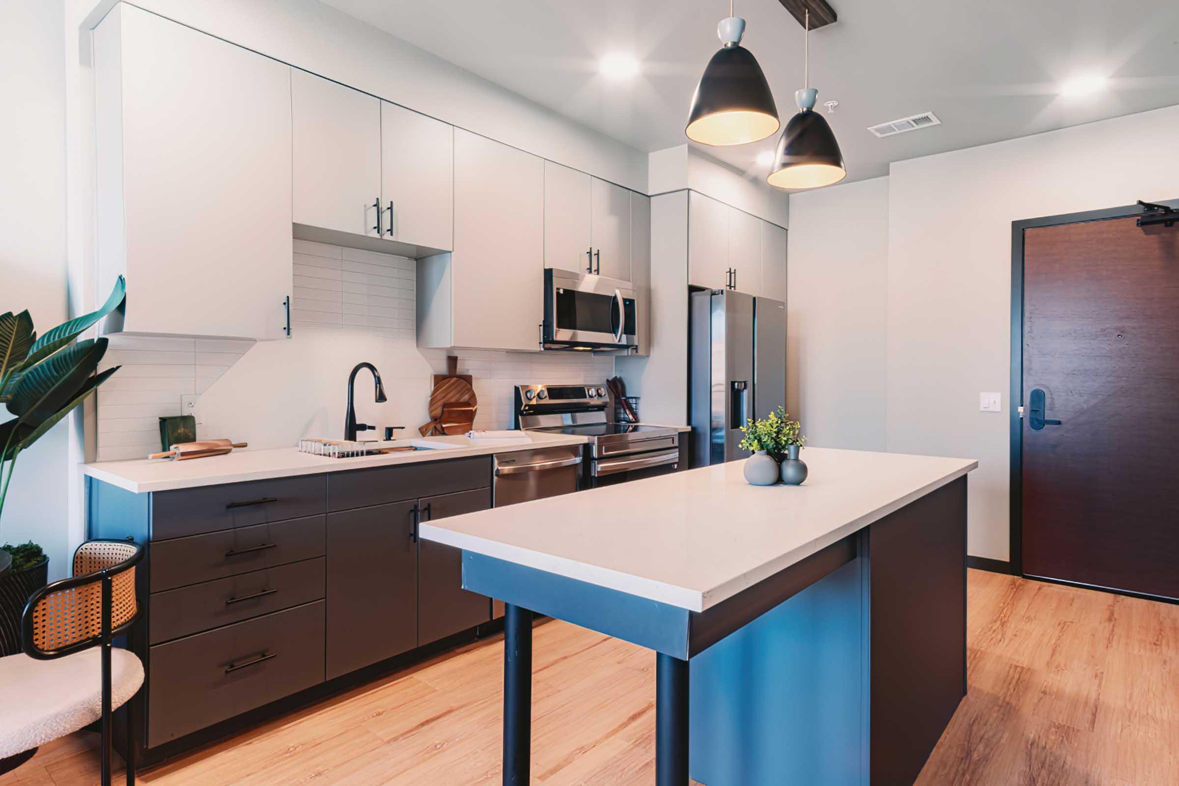 Modern kitchen featuring sleek cabinetry in a blend of light and dark colors, stainless steel appliances, a large island with a minimalist design, and pendant lights hanging above. The space includes a plant and a stylish chair, with wooden flooring adding warmth to the contemporary aesthetic.