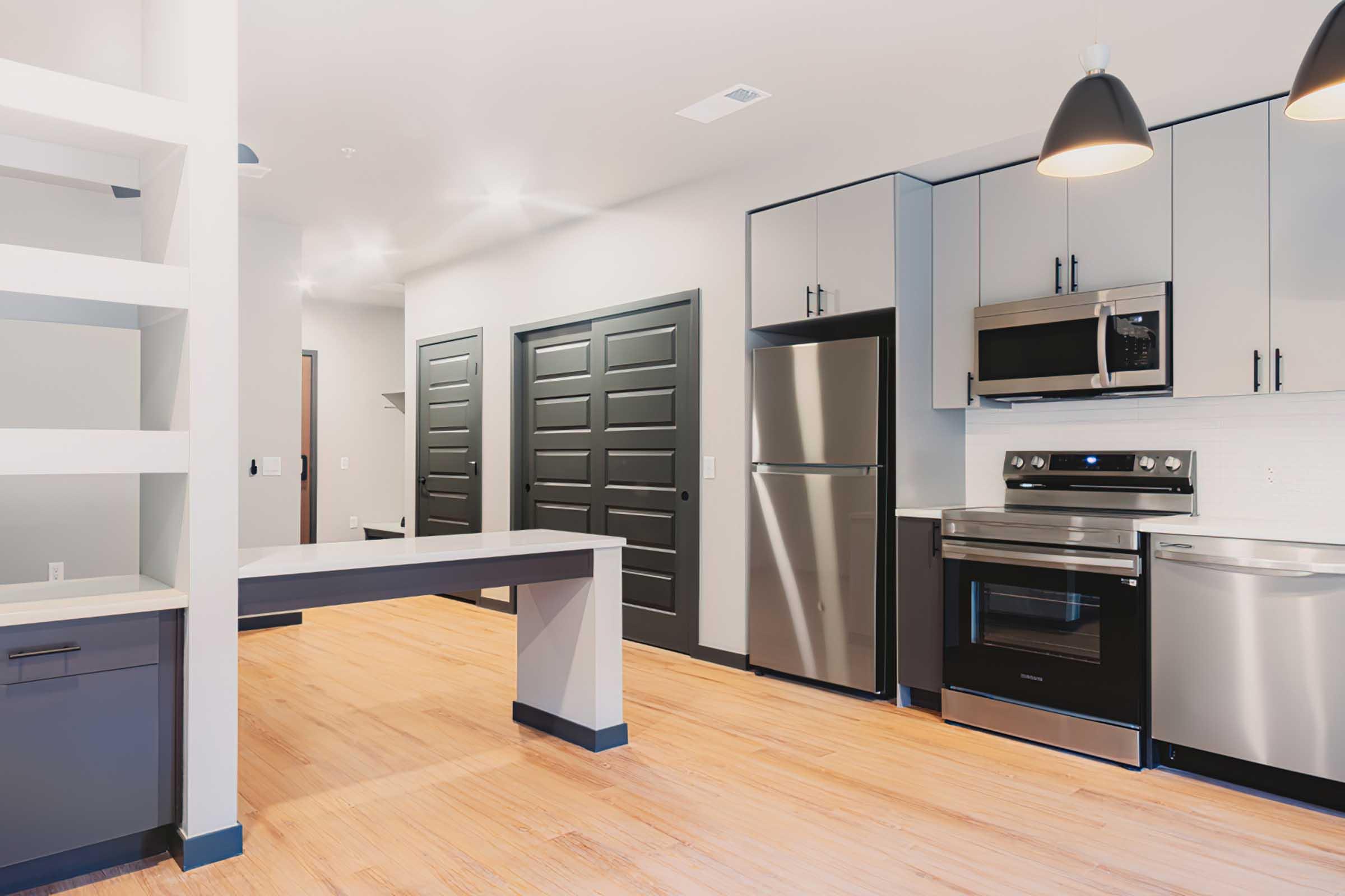 Modern kitchen with stainless steel appliances, including a refrigerator, microwave, and oven. Features sleek cabinetry in gray and black, a light wood floor, and an open layout. Soft lighting from pendant lamps adds a warm ambiance, with a view of a hallway leading to closed doors.