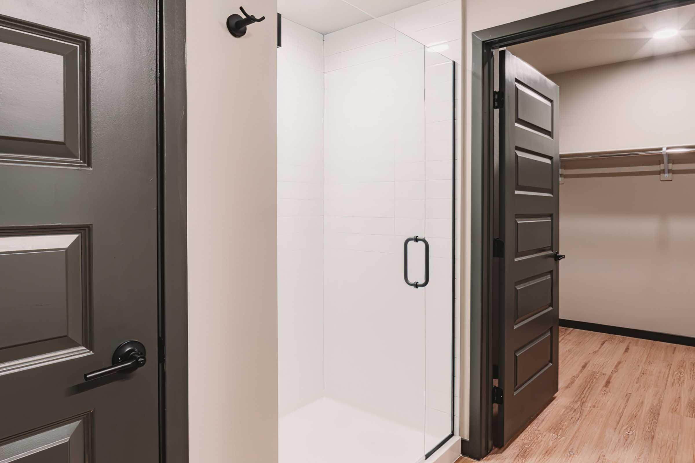 A modern bathroom featuring a clear glass shower enclosure with a black handle. Adjacent to it, there is an open doorway leading to a closet with a hanging rod and wooden flooring. The walls are painted in a neutral tone, and the door is styled in a sleek black finish.