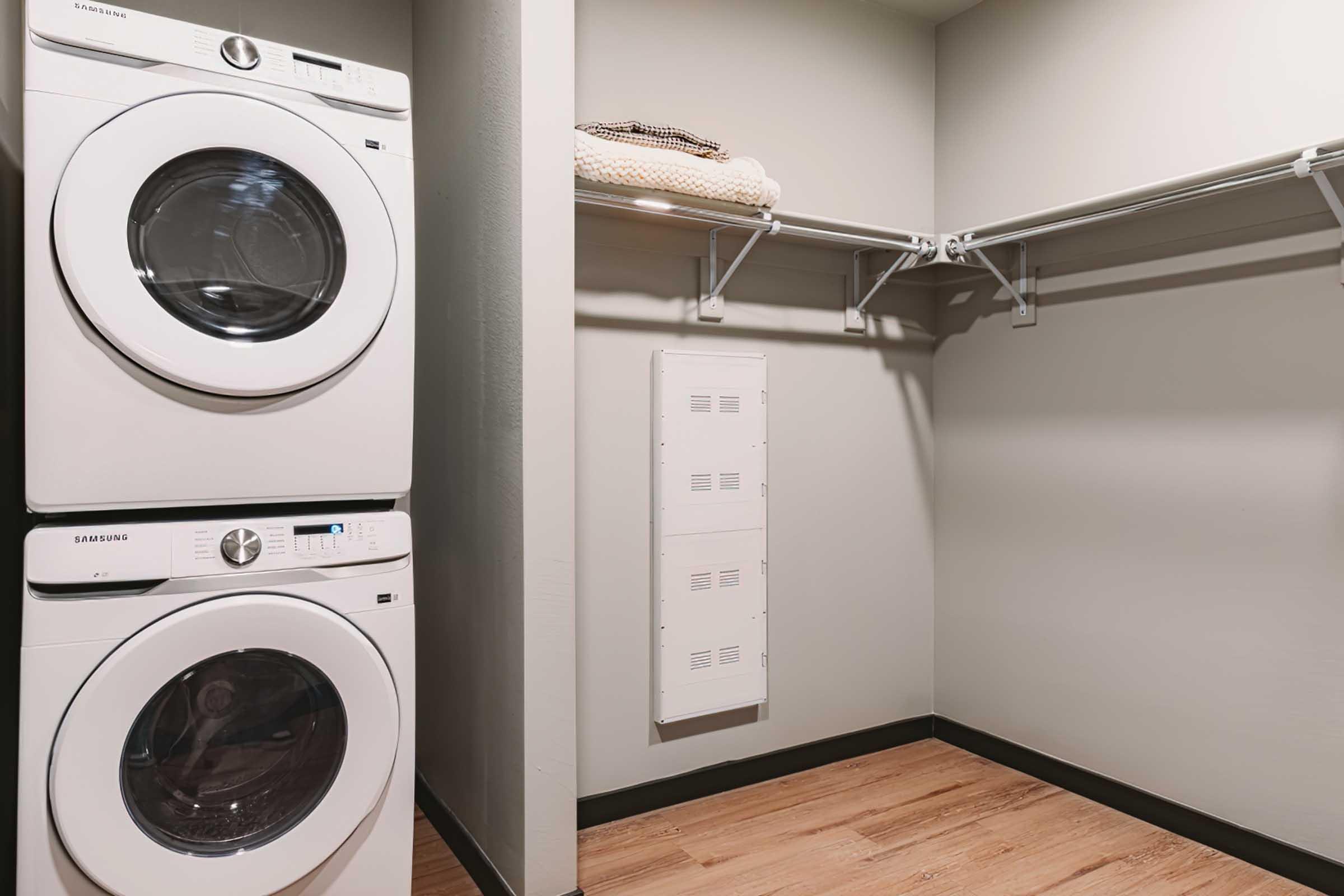A laundry room featuring a stacked washer and dryer in white, positioned next to a bare wall with a shelf for storage. The floor is wooden, and there is a small area for additional storage or utilities on the wall. The overall appearance is modern and clean.