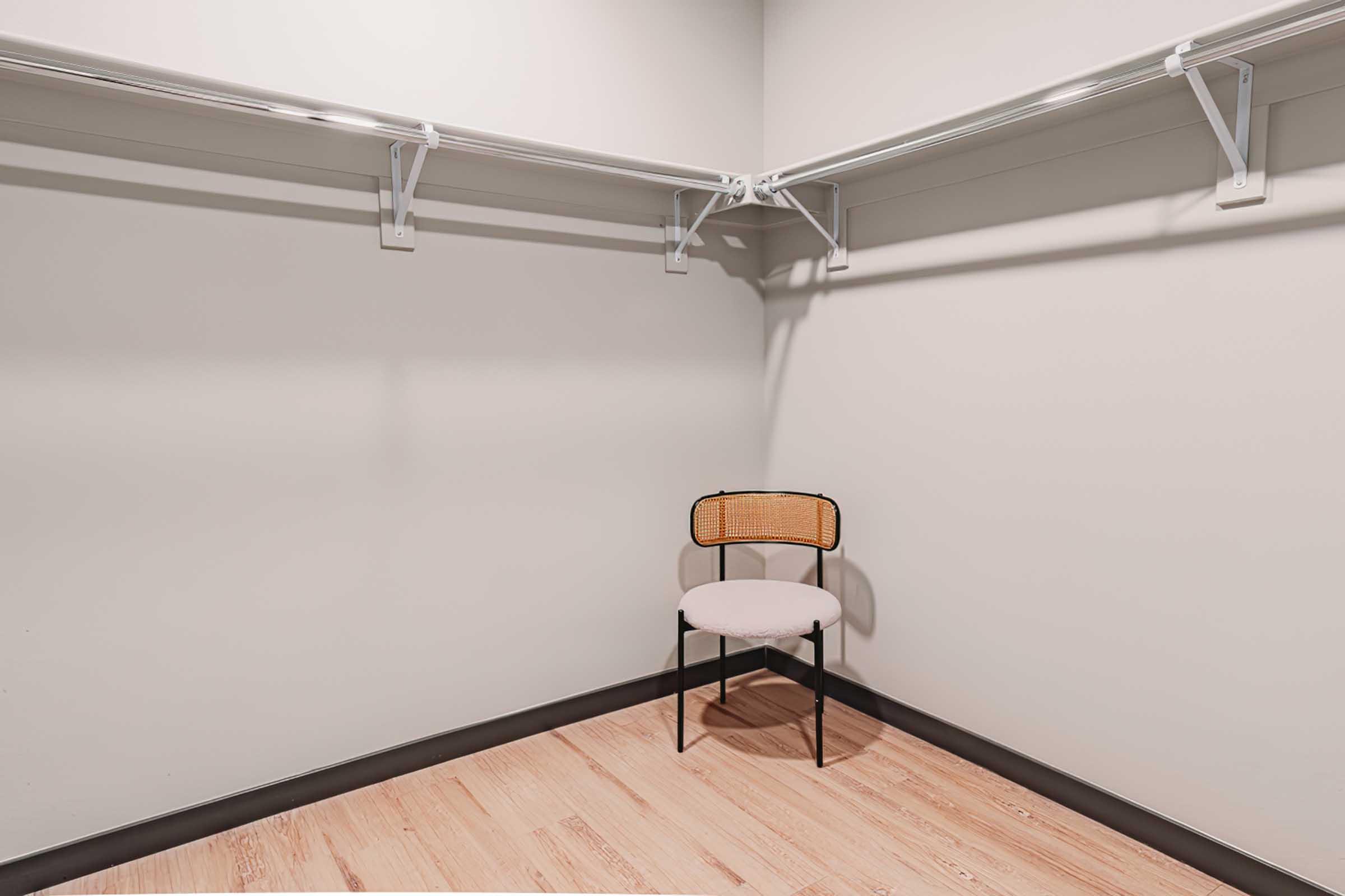 A minimalist empty closet featuring two metal shelving racks on the walls. In the corner, there is a simple chair with a round padded seat and a rattan backrest, set against a light-colored wall and wooden flooring. The space has a clean and uncluttered appearance.