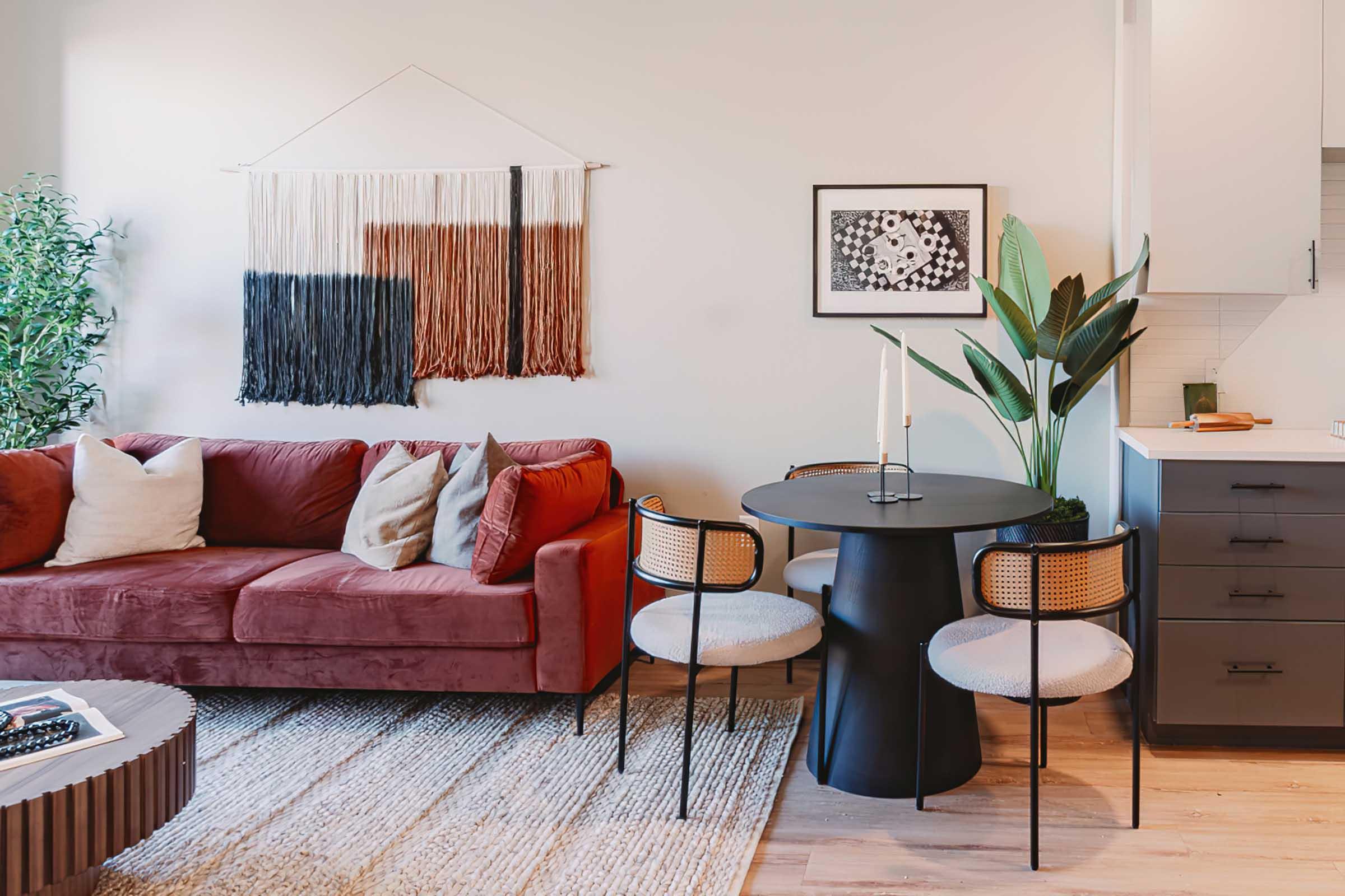 A cozy living space featuring a deep red sofa with decorative pillows, a round black dining table with three stylish chairs, and a modern wall hanging. A green plant adds a touch of nature, and a framed artwork is displayed on the wall, creating an inviting atmosphere.