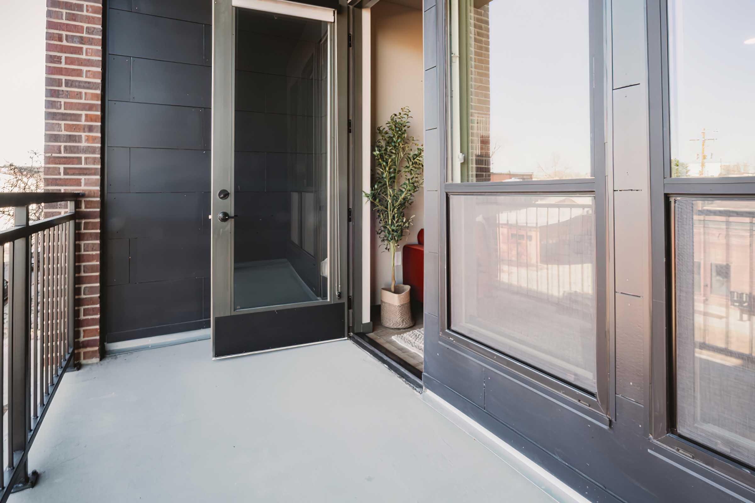 An open balcony door leading to a small outdoor space with a potted plant. The floor is gray, and the walls are black with large windows allowing natural light. A railing surrounds the balcony, creating a modern and inviting atmosphere.