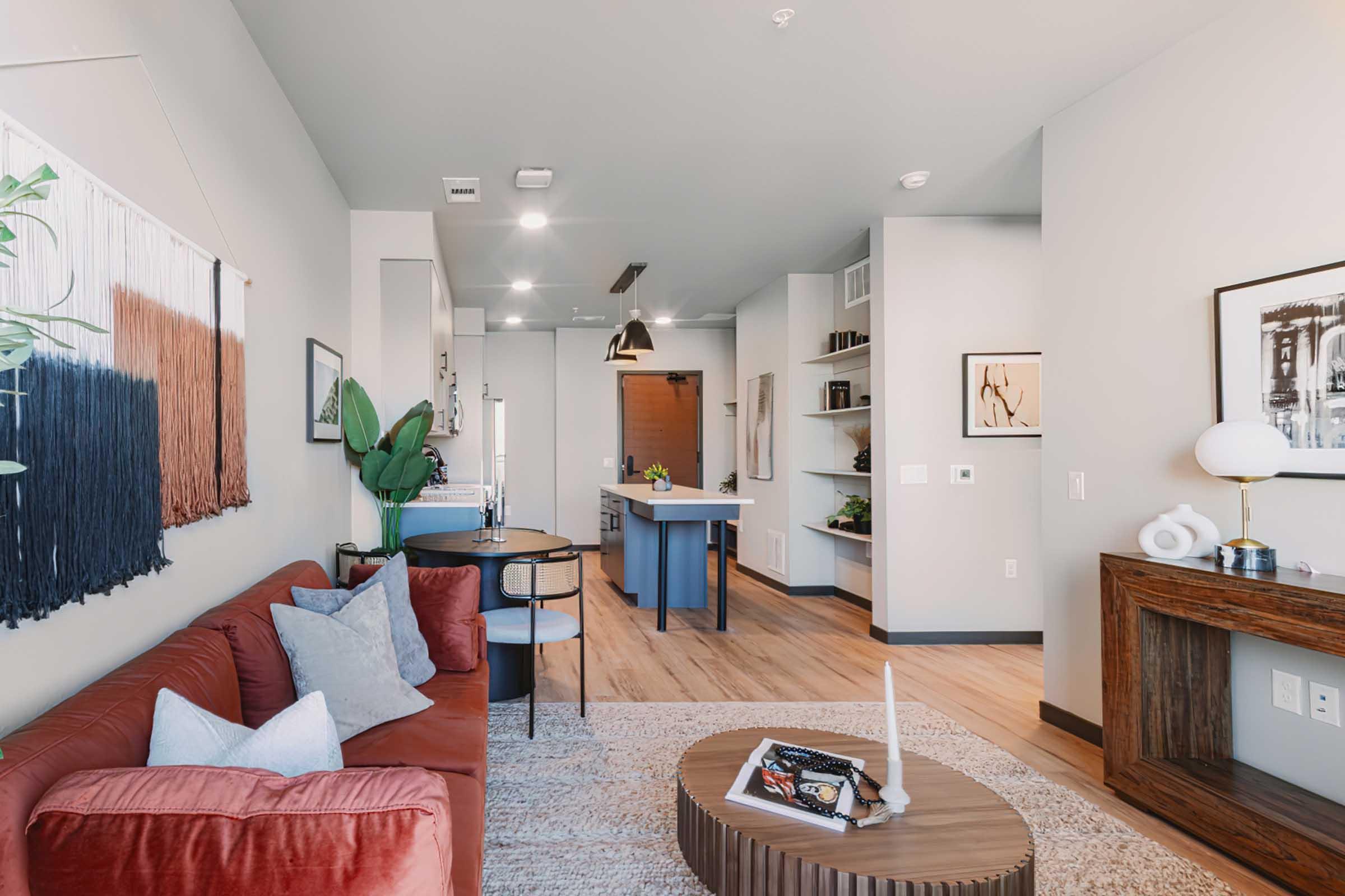A modern living room featuring a red sofa with light cushions, a round coffee table, and a textured rug. There is a plant in the corner, a dining area with chairs and a kitchen island, and a wall with decorative art. Light fills the space from large windows, and wooden flooring adds warmth to the atmosphere.