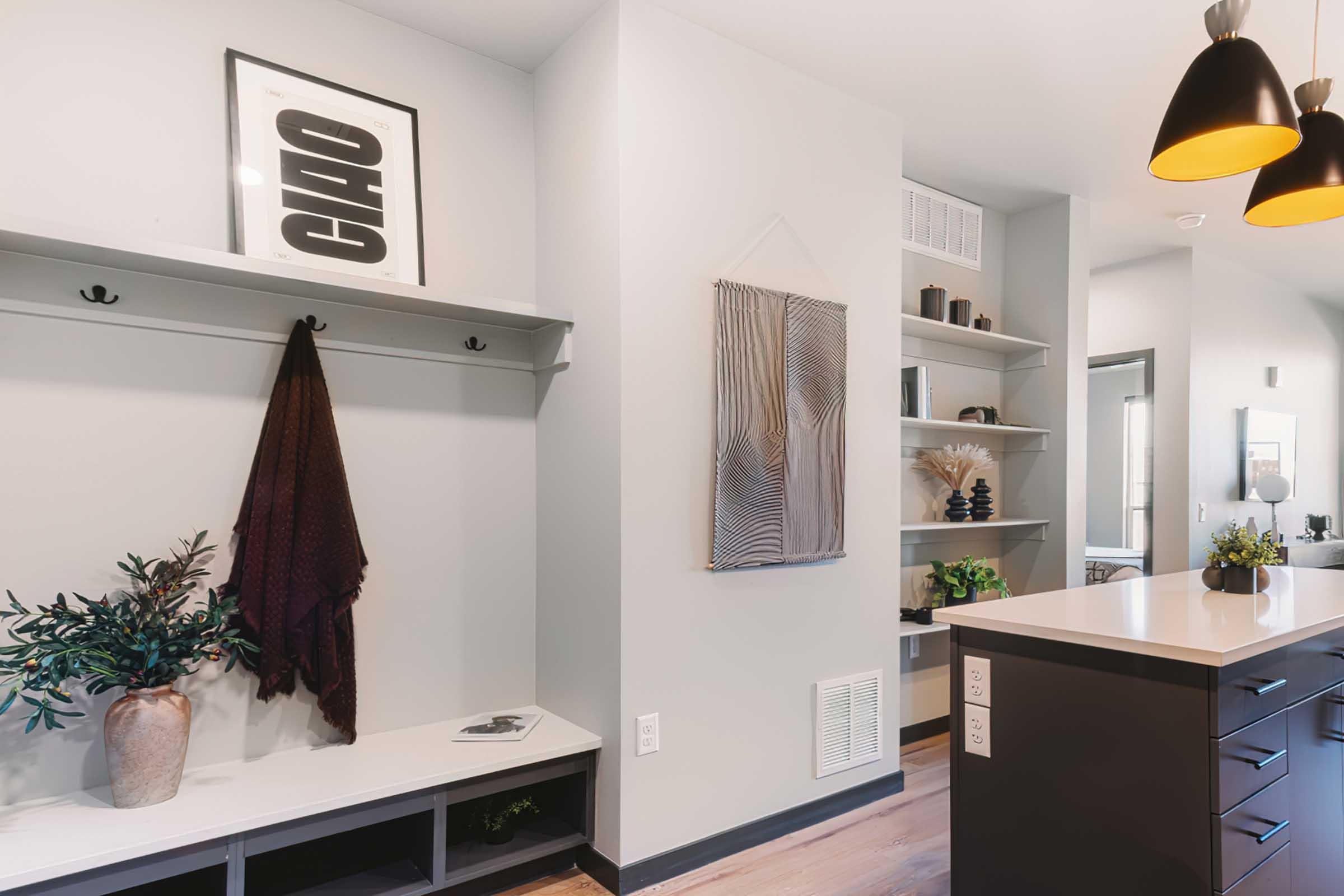Interior view of a modern living space featuring a wall with a framed art piece, a hanging decorative wall piece, and a coat rack. A potted plant and a vase are placed on a bench, while open shelves display decorative items. Soft lighting is provided by pendant lights hanging above a kitchen island.
