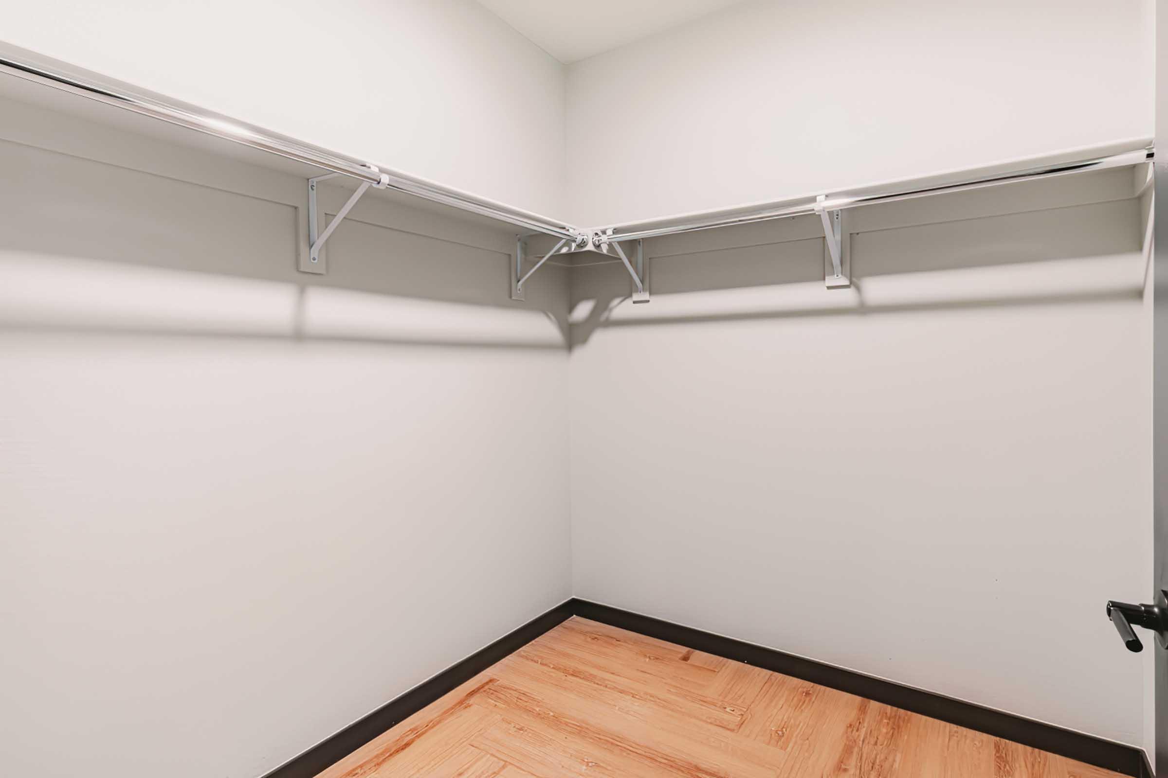 Empty closet with light-colored walls and wooden flooring. The closet features two metal hanging shelves mounted on the walls, providing space for storage. There are no items inside, creating a clean and minimalist appearance.