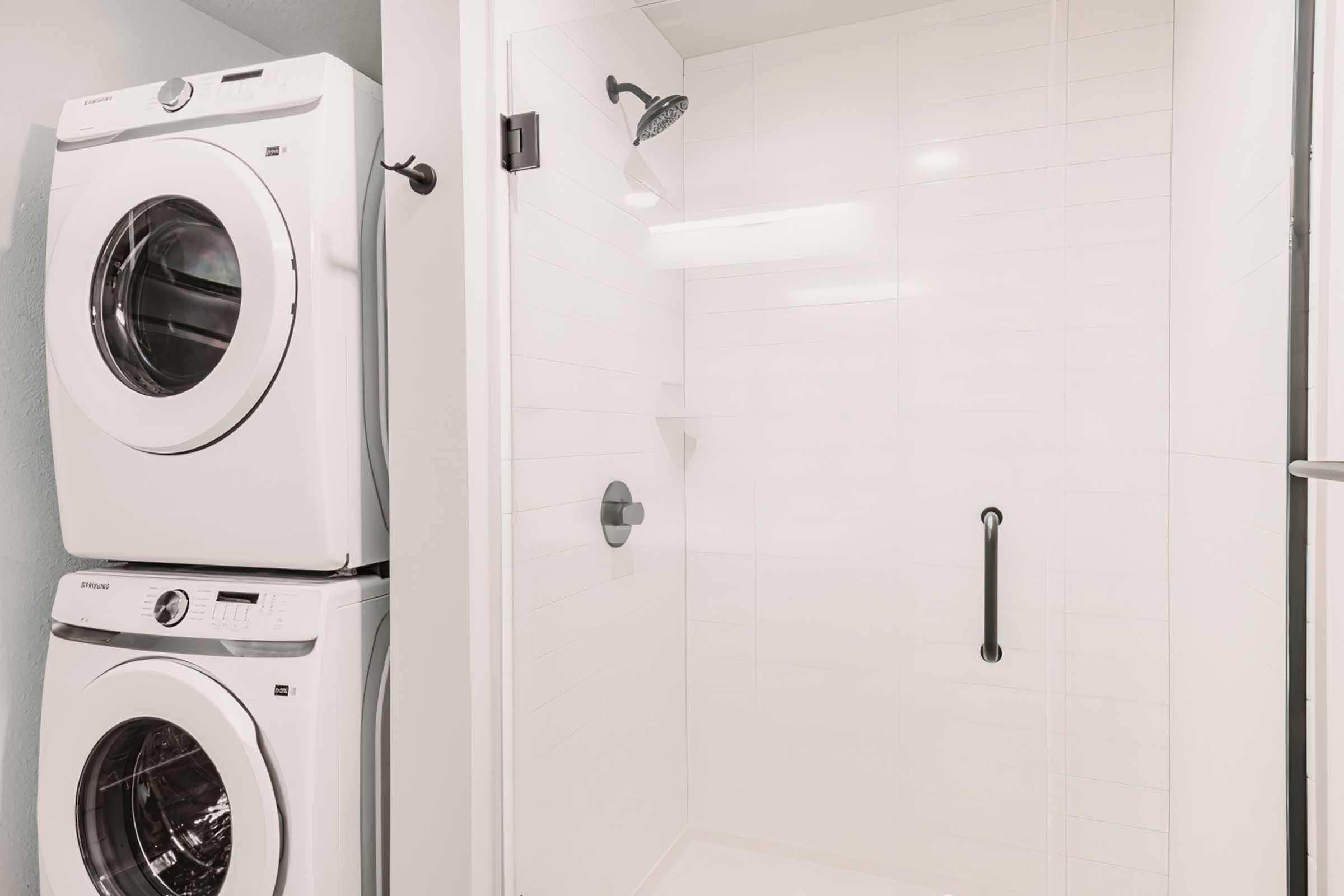 A modern bathroom featuring a stacked washer and dryer to the left and a spacious, minimalist shower with a sleek design, including a handheld shower head and safety grab bar. The walls are tiled in a clean, white finish, contributing to a bright and airy atmosphere.