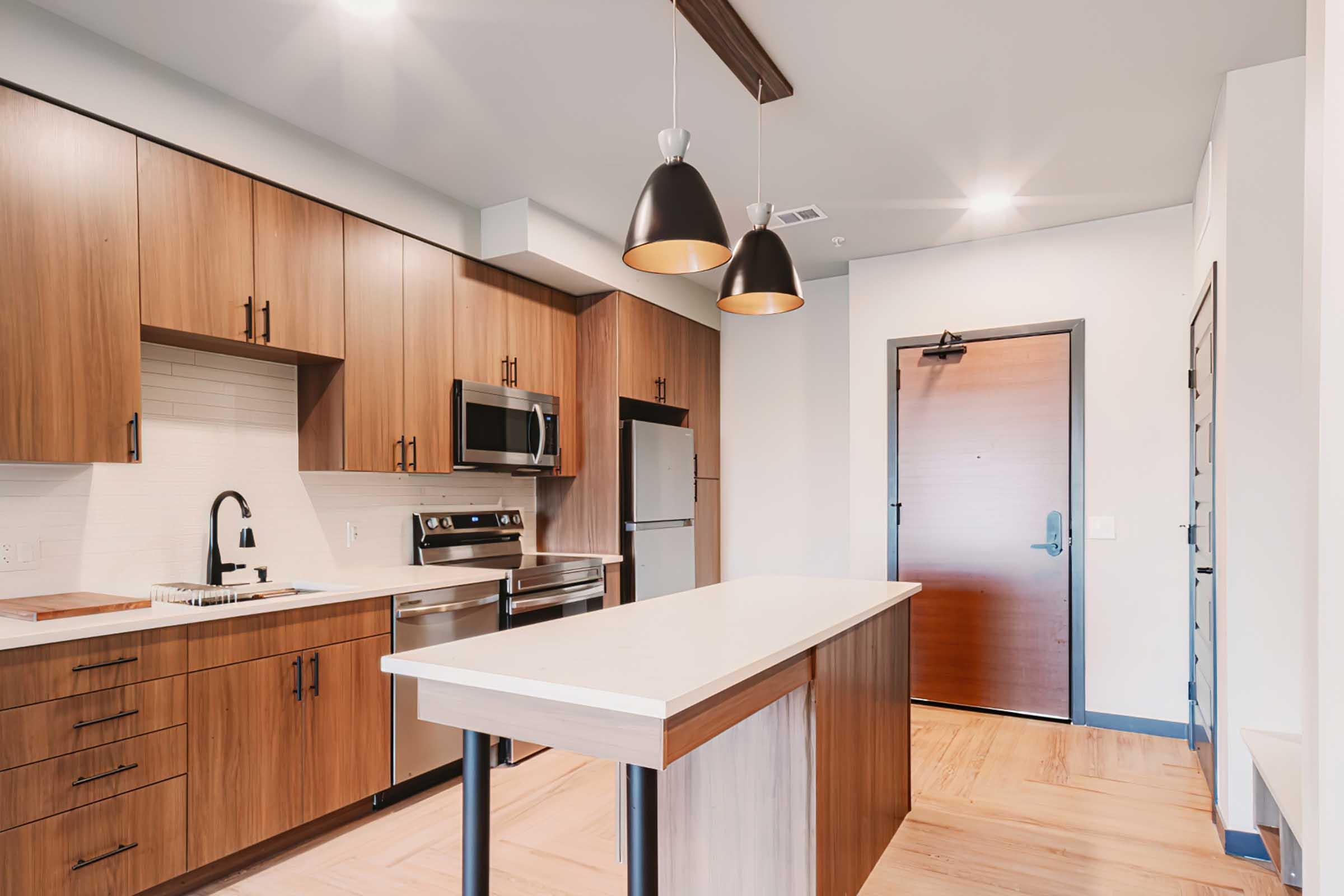 Modern kitchen featuring wooden cabinetry, stainless steel appliances, and a large island with a white countertop. Pendant lights hang above the island, and the space is brightly lit. A wooden door can be seen in the background, leading to the outside.