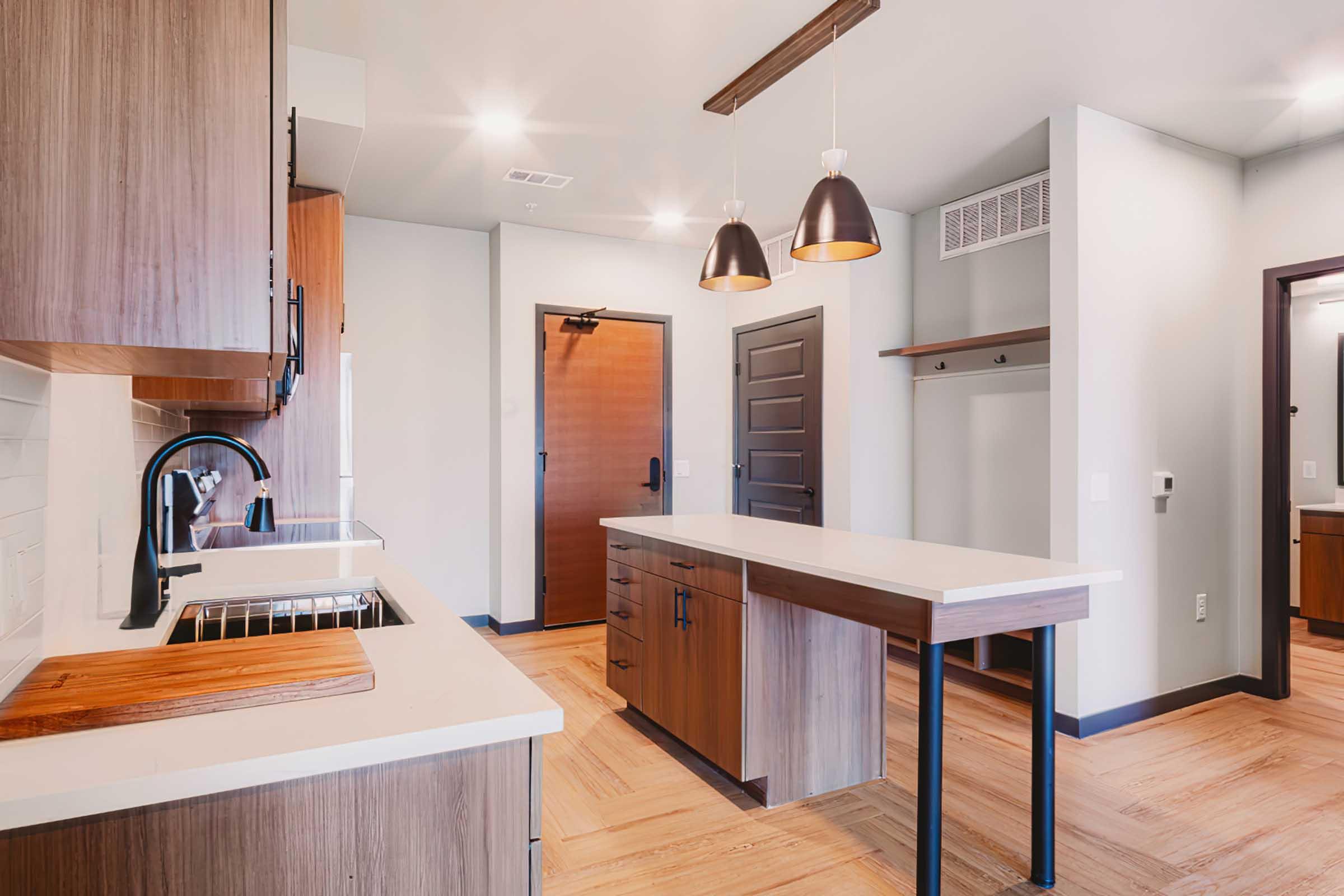 Modern kitchen with wooden cabinetry, a white countertop, and a central island. Two pendant lights hang above the island. The kitchen features a sink and built-in cabinetry, with an adjacent area that includes a door and hooks for hanging items. The floor is made of wooden planks, creating a warm ambiance.