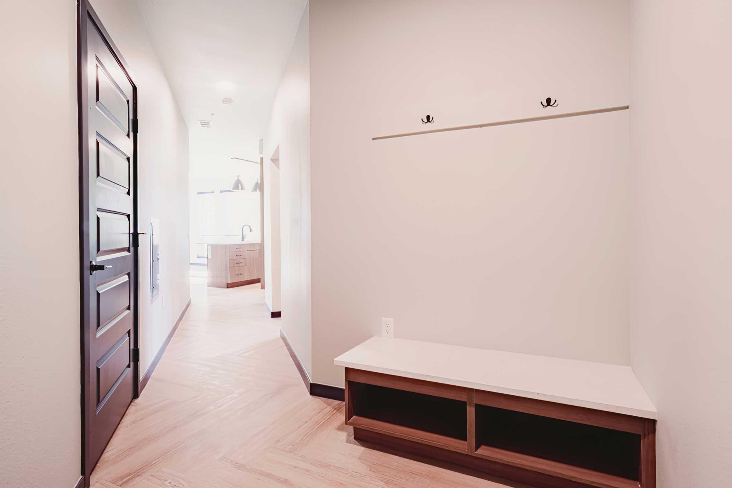 A narrow hallway featuring light-colored walls and wooden flooring. On one side is a simple wooden bench with a white top and open storage below, accompanied by two wall-mounted hooks above. The hallway leads to a well-lit area, suggesting a kitchen or living space in the background.