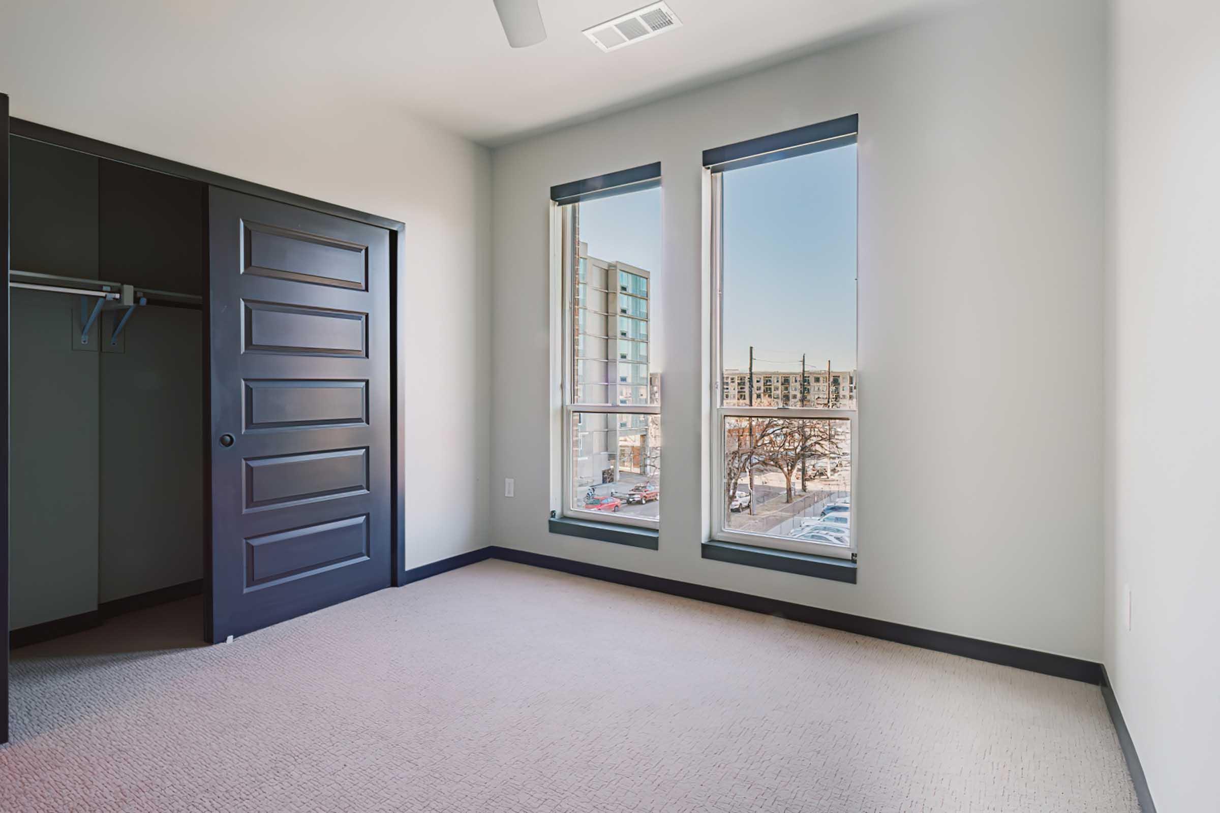 Empty bedroom with beige carpet, gray walls, and a large window featuring a view of exterior buildings. A closet with a dark door is visible on the left. Natural light enters through the windows, creating a bright and airy atmosphere.
