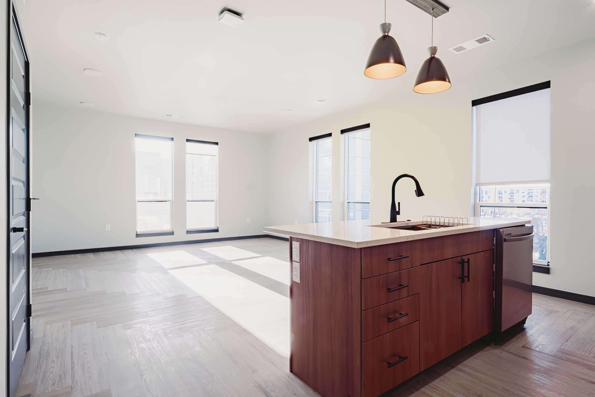 Modern, open-concept kitchen and living area featuring a central island with a dark wood finish, a black faucet, and pendant lighting. Large windows allow natural light to fill the space, complemented by light-colored walls and flooring. Minimalist design elements create a contemporary ambiance.