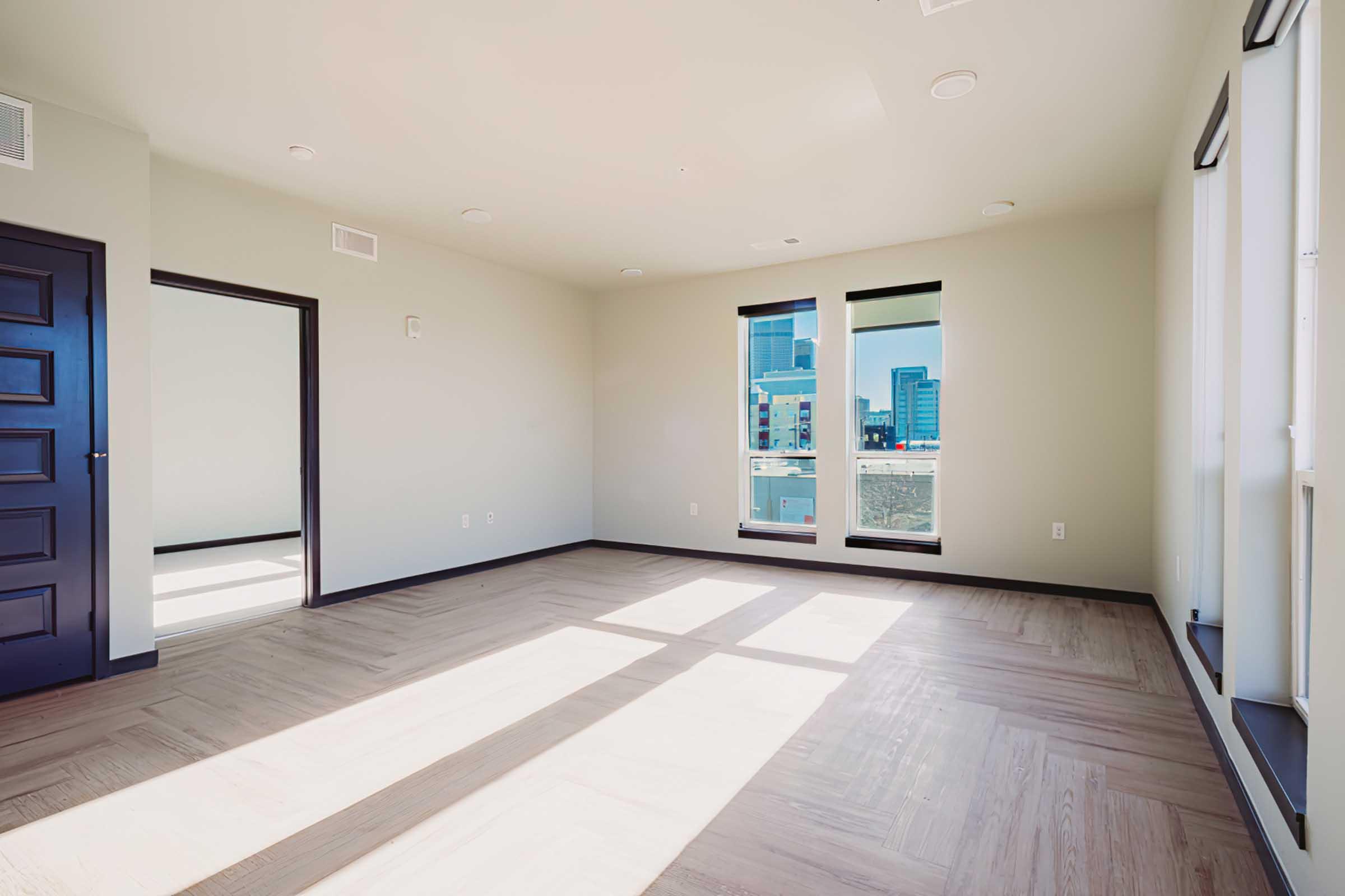 A spacious, empty room with hardwood flooring and large windows allowing natural light to fill the space. There are two windows on the left side, and a dark-colored door on the right leads to another area. The walls are painted in a neutral color, enhancing the bright ambiance of the room.