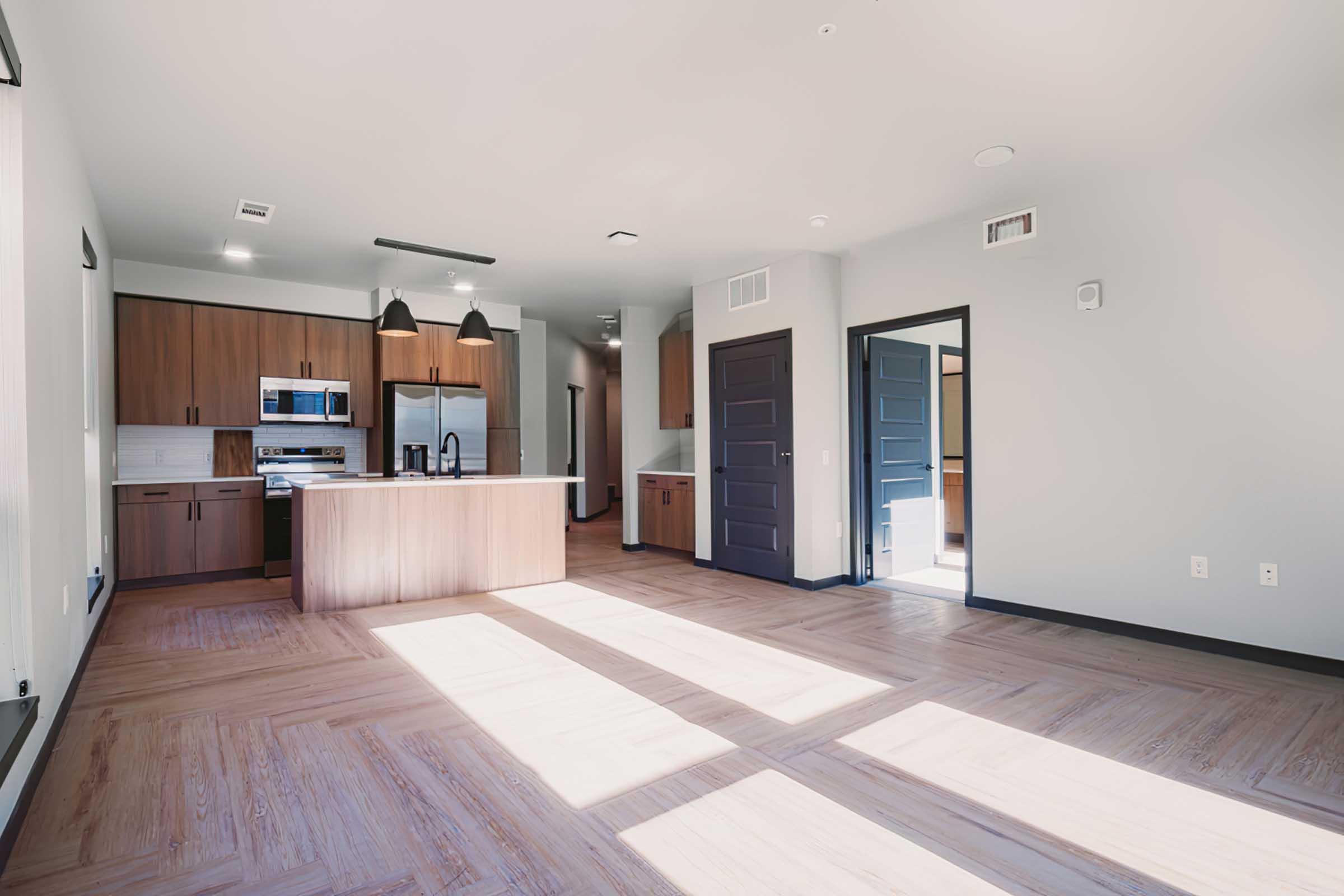A modern open-concept living space featuring a kitchen with stainless steel appliances, wooden cabinets, and a large island. The room has ample natural light streaming in through windows, highlighting the sleek flooring and minimalist design. The walls are painted in neutral tones, and doors are dark.