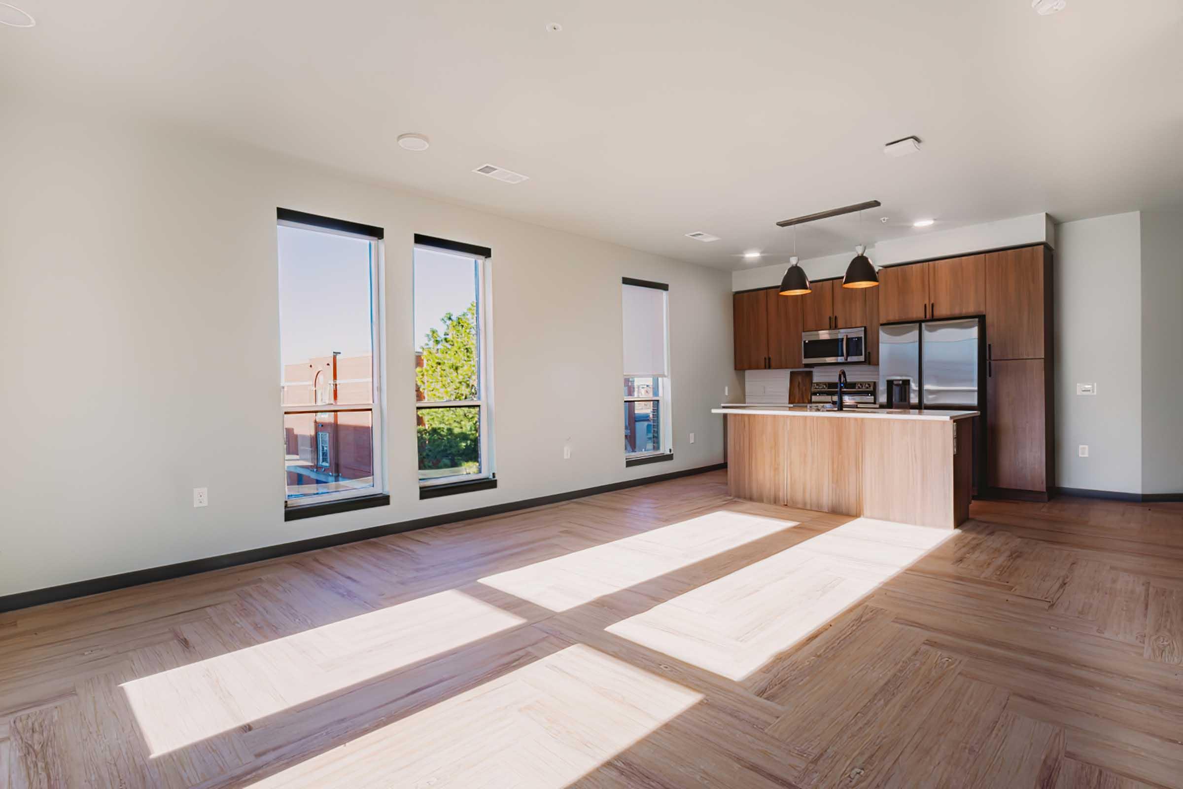 A modern, spacious living area featuring large windows that allow natural light to fill the room. The space includes a kitchen with dark wood cabinets, stainless steel appliances, and two pendant lights hanging above the kitchen island. The flooring is light wood, creating a cozy and inviting atmosphere.