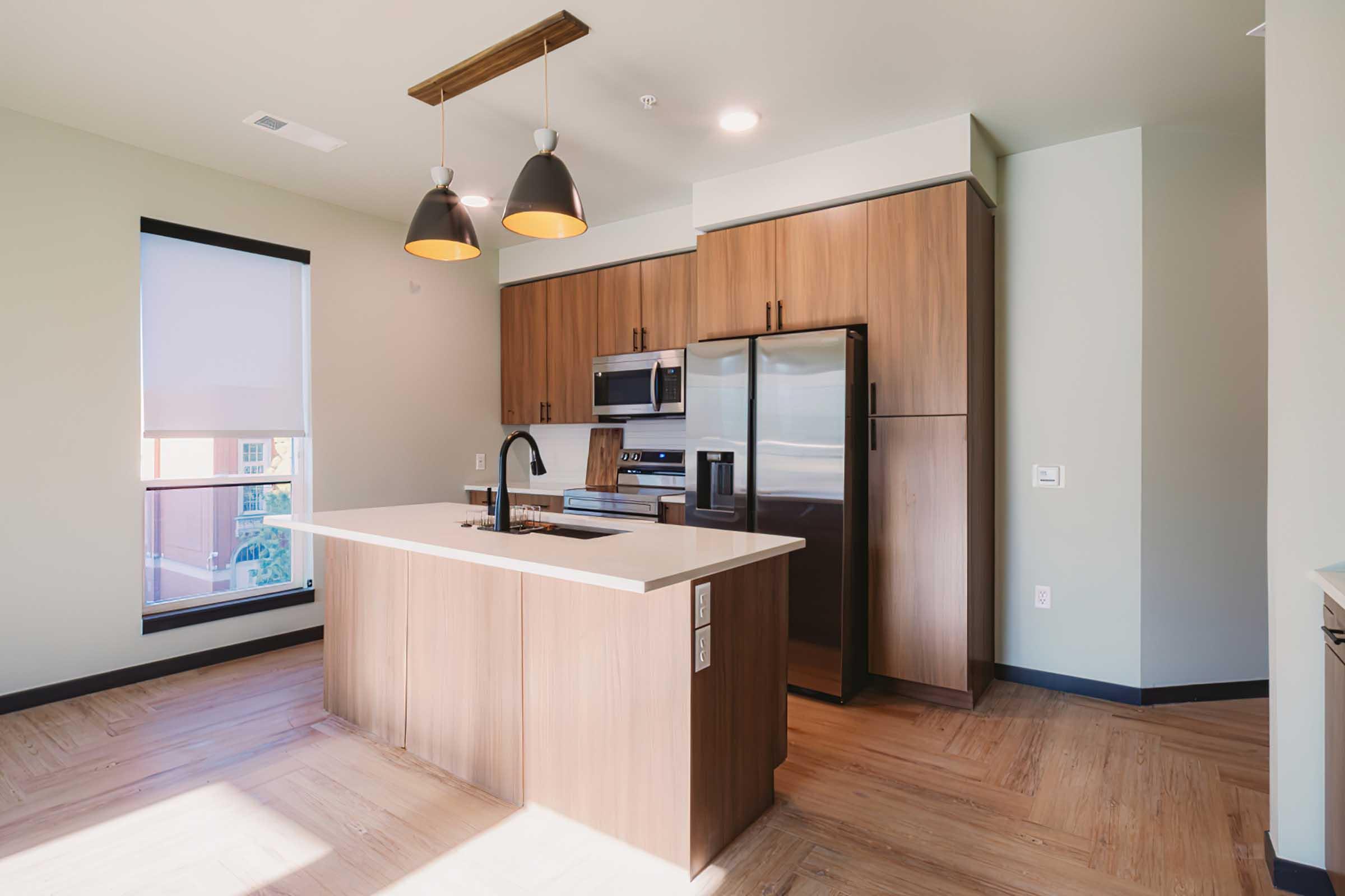 Modern kitchen with wooden cabinetry, stainless steel appliances, and a large island. The space features pendant lighting above the island and ample natural light from a nearby window. The flooring is a light-hued wood, creating a warm and inviting atmosphere.