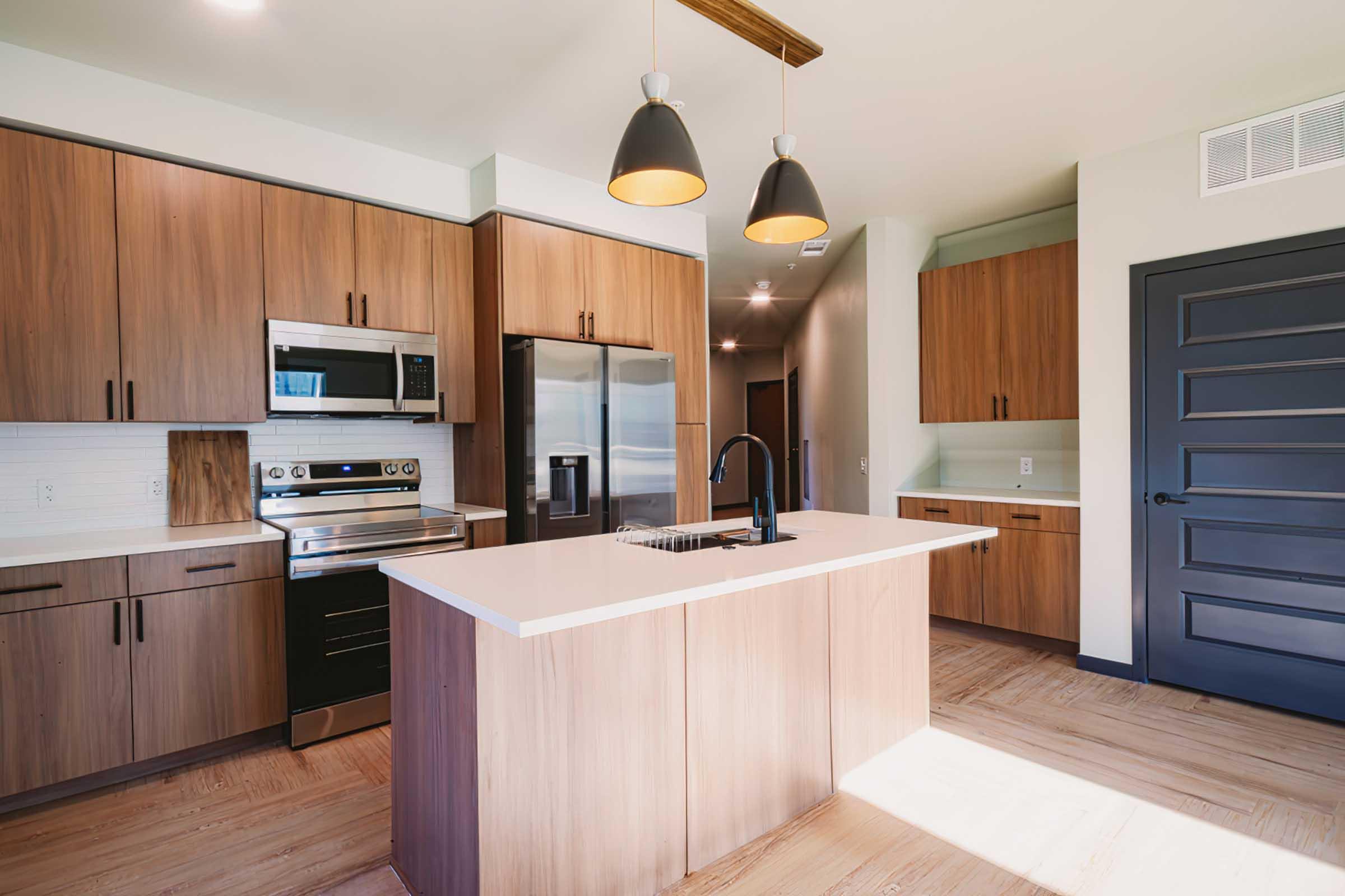 Modern kitchen with wooden cabinetry and stainless steel appliances. It features a central island with a sink, stylish pendant lighting, and a dark door contrasting with the light walls. The space has a warm natural wood floor that enhances the contemporary design.