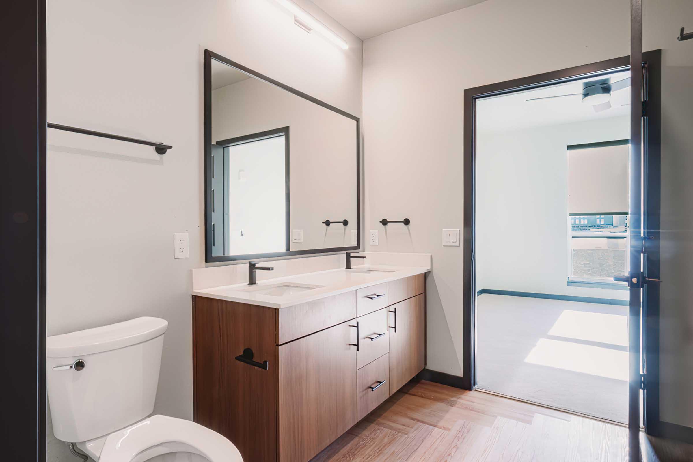 A modern bathroom featuring a wooden vanity with dual sinks, a large mirror, and a sleek black faucet. A white toilet is positioned to the left, and a doorway on the right leads to a well-lit room with a window. The floor is wooden, and the walls are painted light gray.