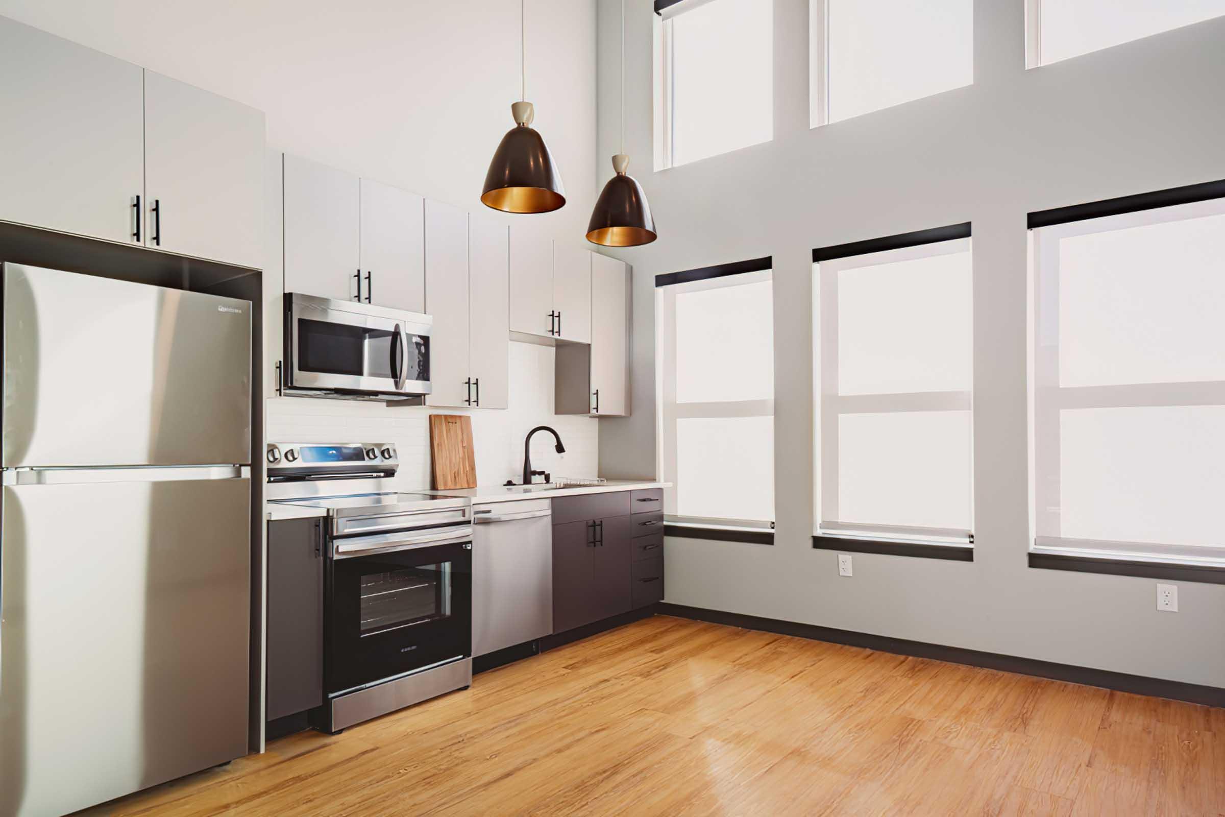 Modern kitchen featuring stainless steel appliances, including a refrigerator, microwave, and oven. The sleek cabinetry is a mix of light and dark tones. Large windows allow natural light to fill the space, complemented by stylish pendant lighting overhead. The floor is made of warm wood, enhancing the contemporary aesthetic.
