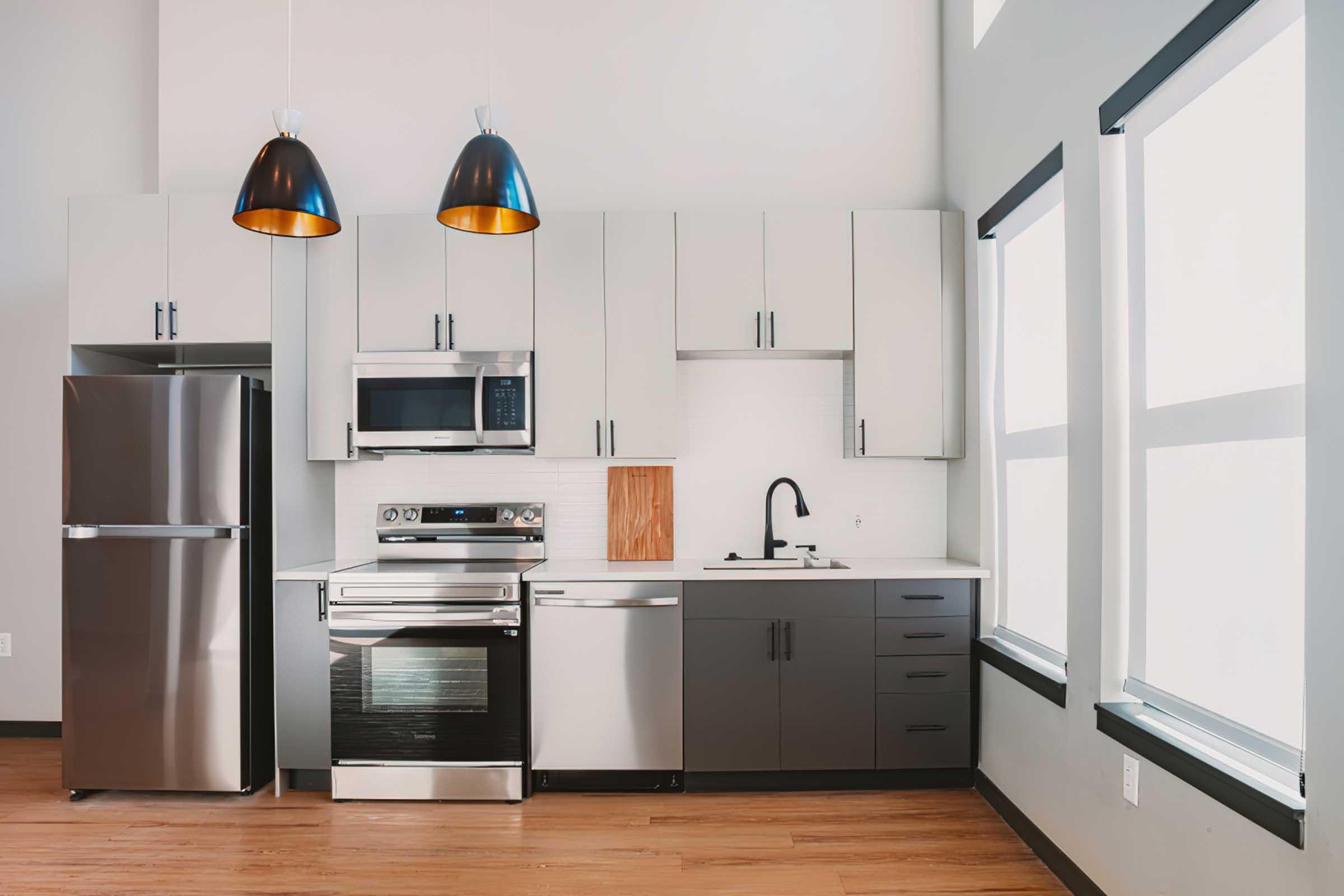 Modern kitchen featuring stainless steel appliances including a refrigerator, microwave, oven, and dishwasher. The cabinets are a mix of light and dark colors. Two pendant lights hang above the countertop, and large windows allow natural light to fill the space, enhancing its contemporary design.
