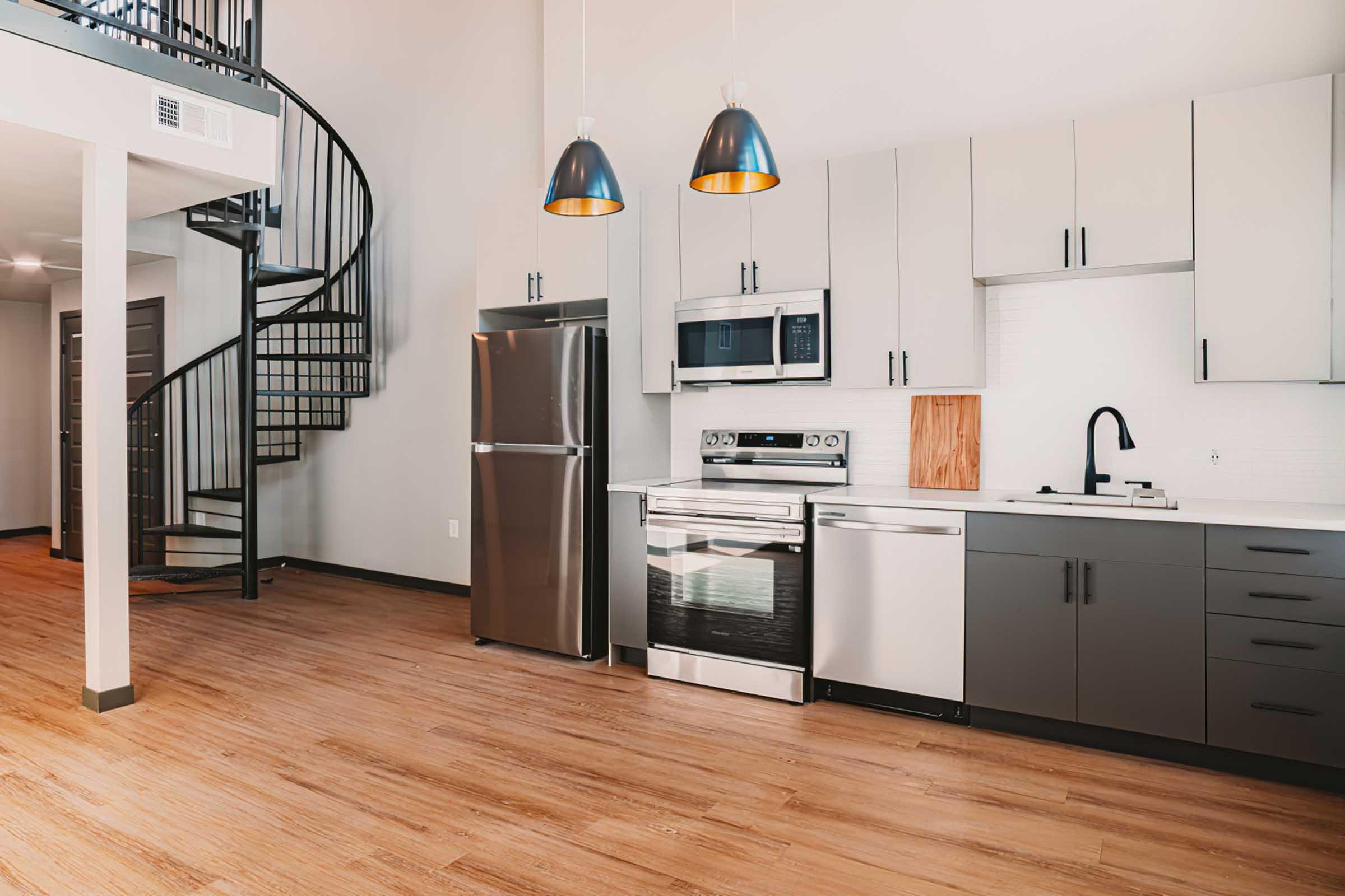 A modern kitchen featuring stainless steel appliances including a refrigerator, microwave, and oven. The design includes sleek cabinetry, a black sink, and a wooden cutting board. A spiral staircase is visible in the background, and the floor is finished with light wood.