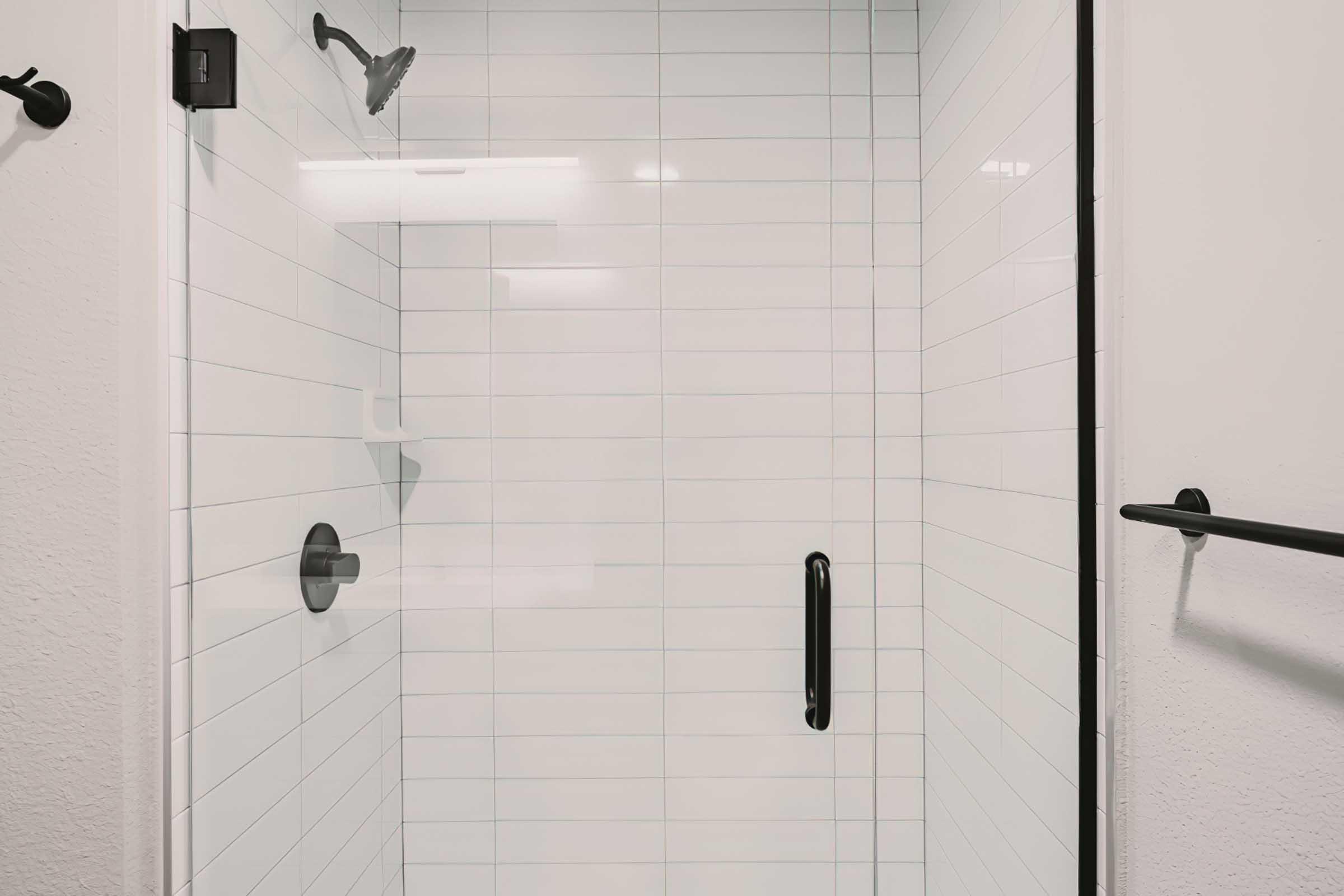 A clean and modern shower stall featuring white tiled walls, a glass door, a sleek black handle, and minimalistic shower fixtures. The space is brightly lit, with a simple design that emphasizes cleanliness and contemporary style.