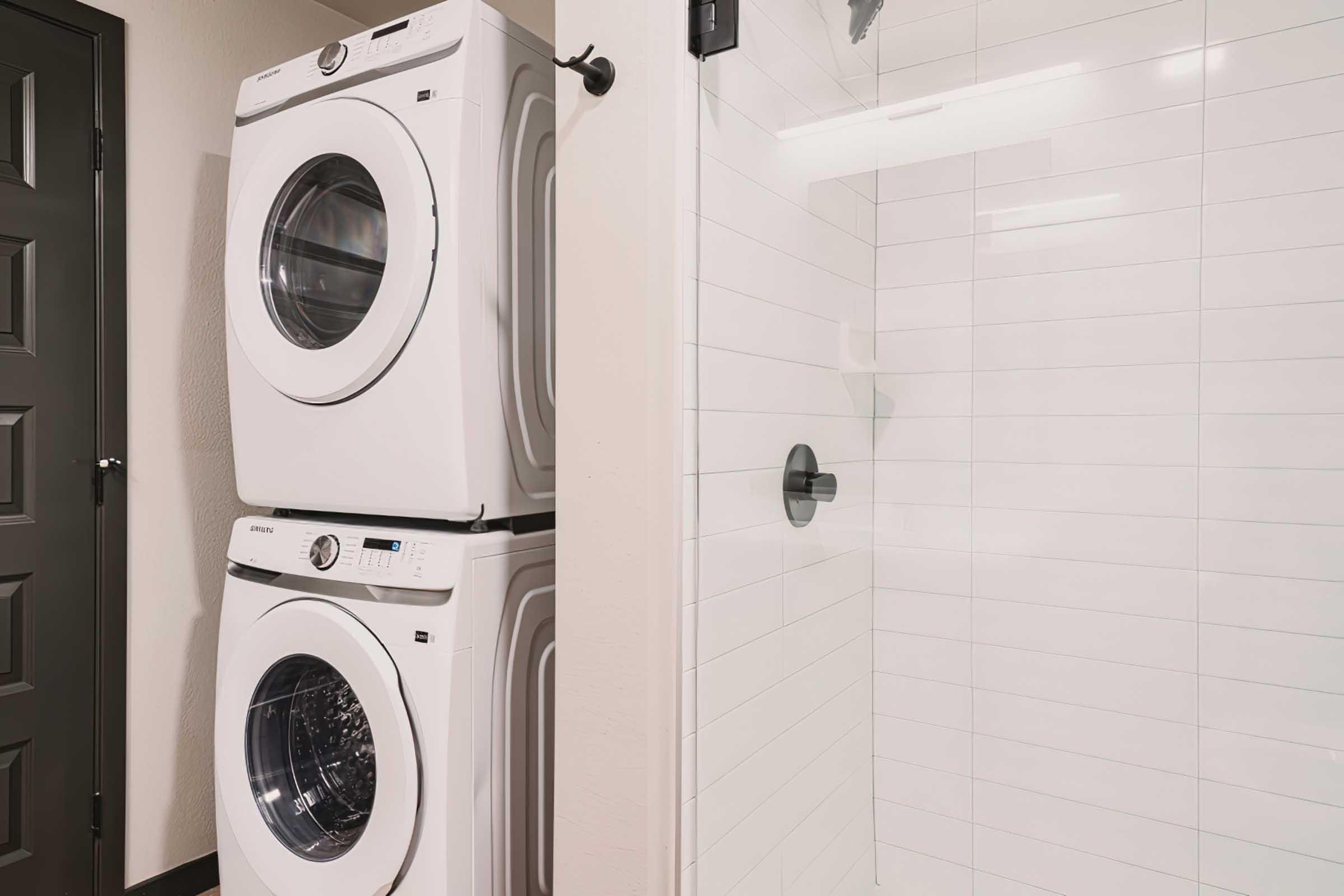 A modern laundry area featuring a stacked washer and dryer unit in white, next to a glass-enclosed shower with white tile walls. The background includes a dark door, providing a contrast to the light-colored appliances and fixtures. The space has a clean and contemporary design.