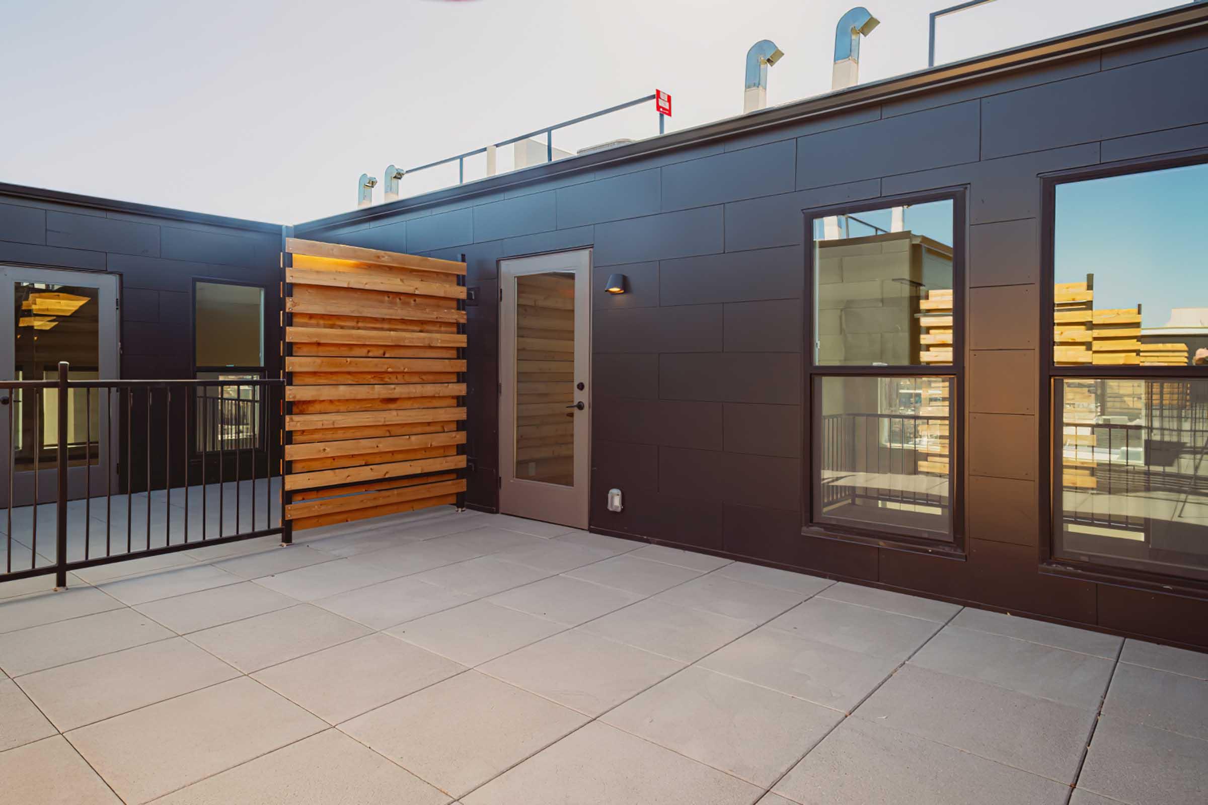 A modern rooftop terrace featuring sleek black walls, large windows reflecting the surroundings, and a wooden slatted wall. The space is equipped with a metal railing and tiled flooring, offering a contemporary outdoor area for relaxation or entertainment.