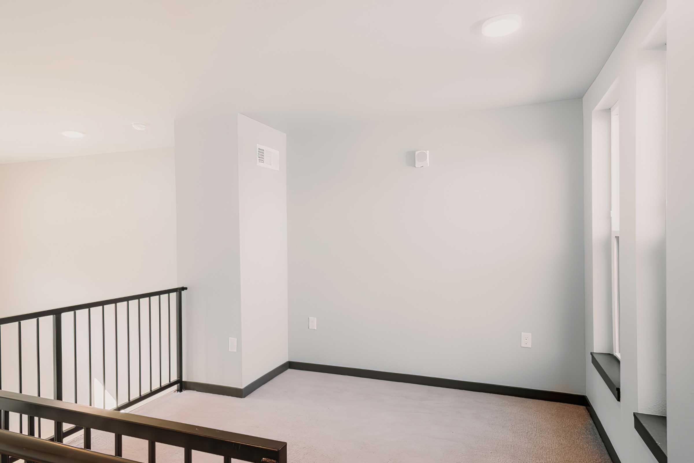 Empty interior space featuring light-colored walls and a carpeted floor. A black railing lines a staircase, leading to a well-lit area with two windows. The room has a minimalist design and appears to be recently renovated or new, creating a bright and airy atmosphere.