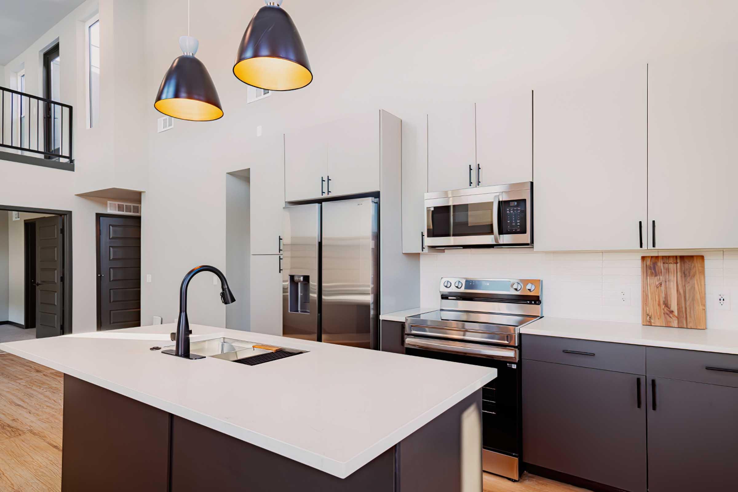 Modern kitchen featuring sleek dark cabinetry and stainless steel appliances. A large island with a built-in sink sits at the center, complemented by industrial-style pendant lights. The space has a spacious, open layout with natural light coming in from large windows.